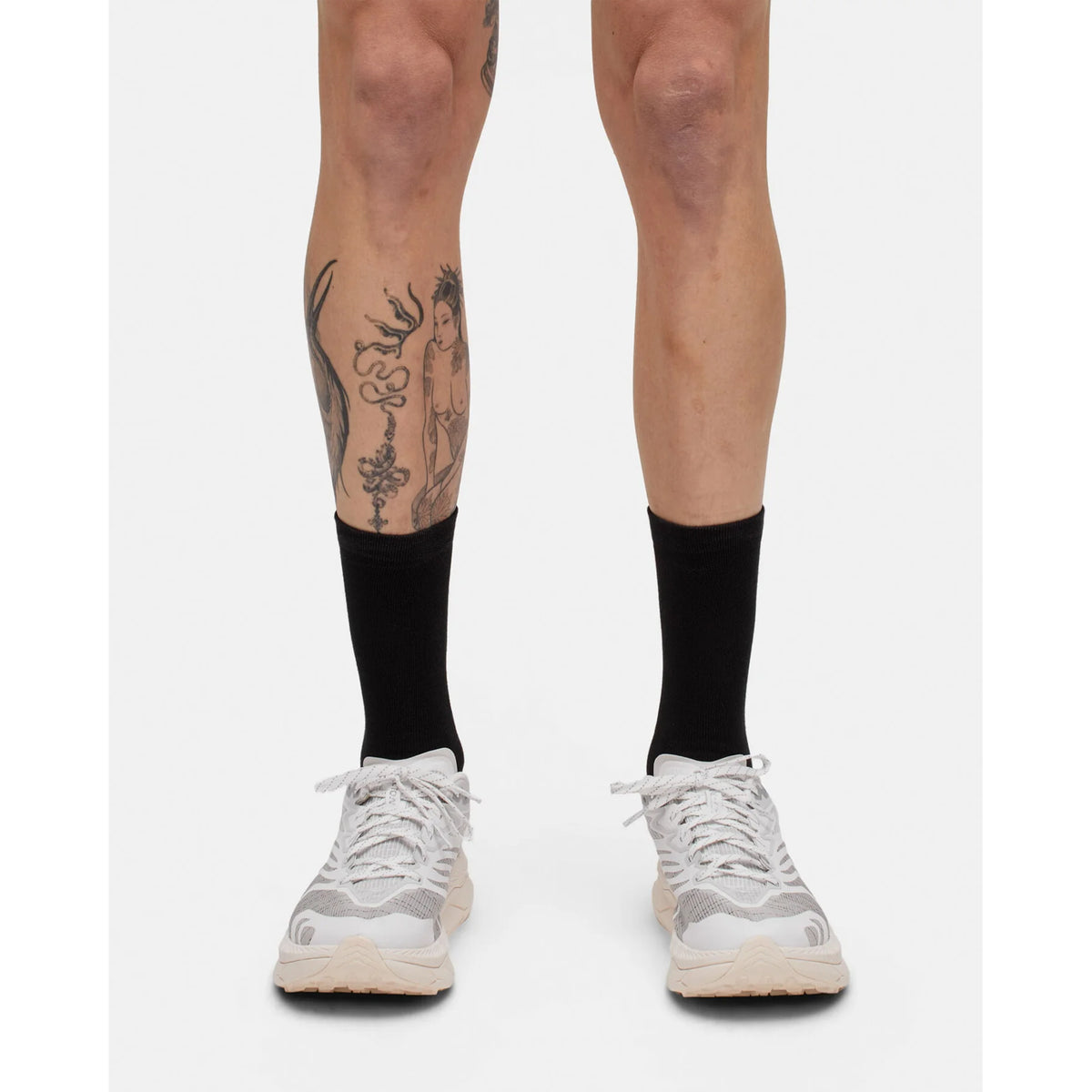 A person wearing Portal Pattern Active Socks in Black Beauty / Reflective—which feature moisture control—paired with white sneakers, displays leg tattoos while standing against a plain white background. Only their lower legs and feet are visible.
