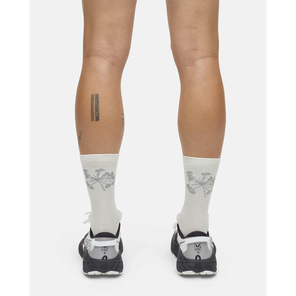 A person is standing, viewed from behind, wearing Portal Pattern Active Socks Bone White—lightweight synthetic socks designed for moisture control during activity. The socks feature a gray palm tree design and are paired with black-and-white athletic shoes. The person has two tattoos on their calves: one with vertical lines and another with script. The background is plain white.