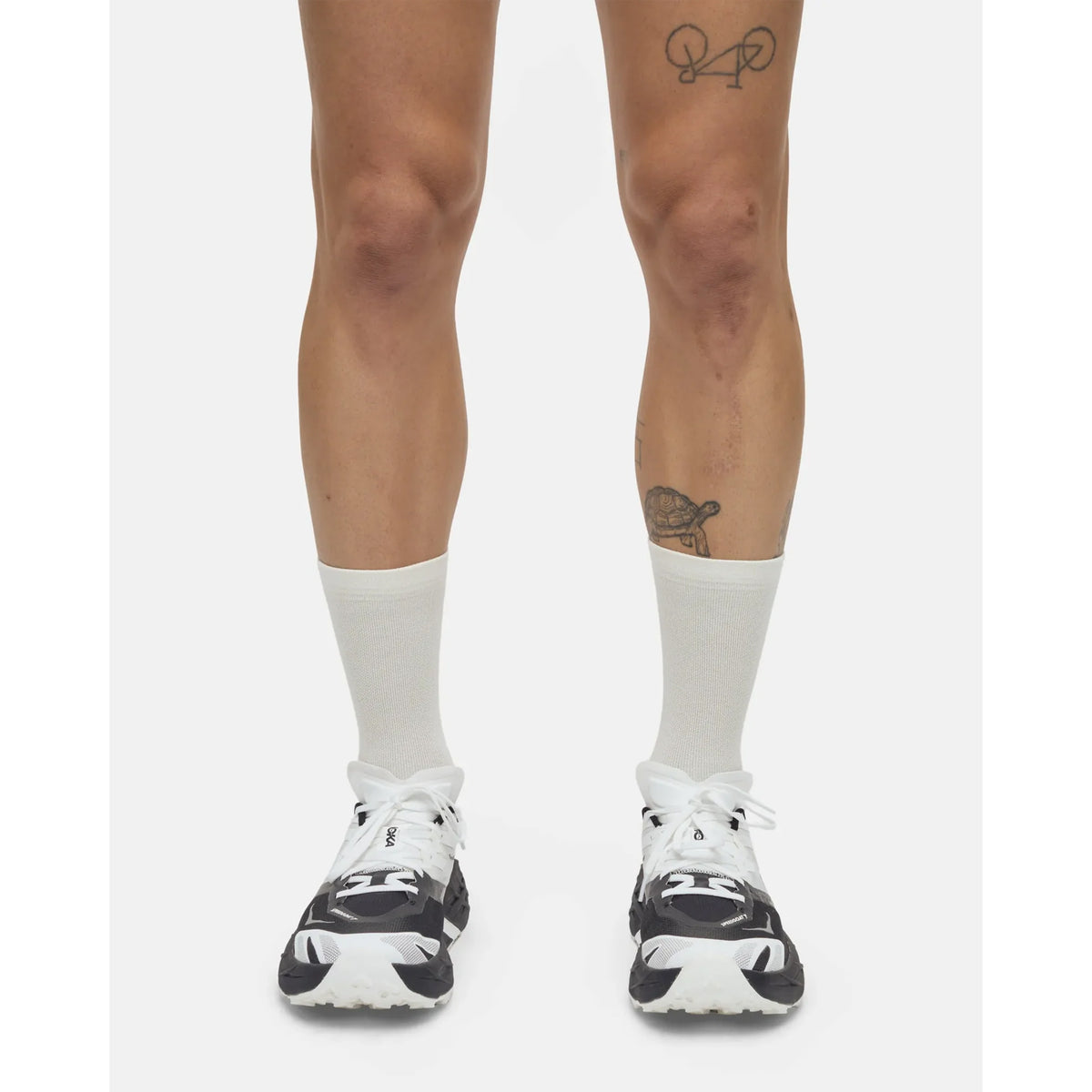 A person stands with legs visible from the knees down, wearing Portal Pattern Active Socks in Bone White and black-and-white sneakers. Two tattoos are visible: a bicycle on the left thigh and another on the right shin. The background is plain white.