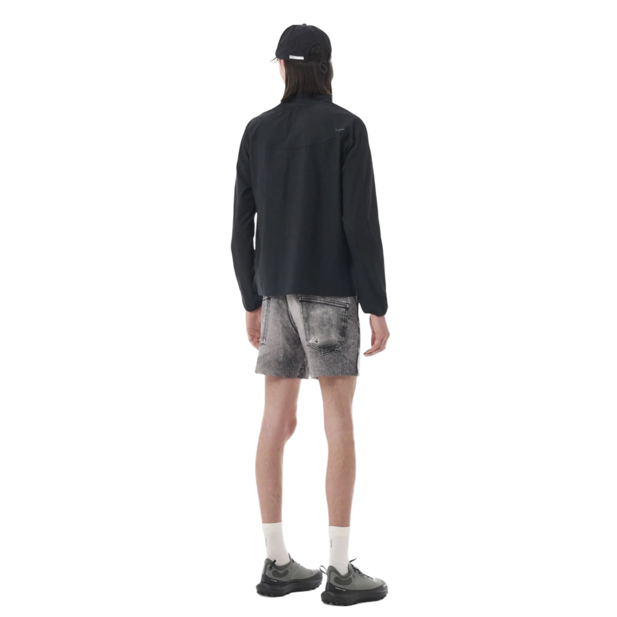 A person is shown standing with their back to the camera, wearing a black Satisfy PeaceShell River Shirt—a technical running shirt designed for lightweight wind protection—paired with a black cap, black and gray shorts, white socks, and black shoes. The background is plain white.