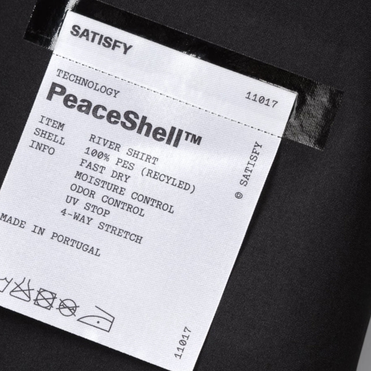 Close-up of a white clothing label on dark fabric. The label highlights Satisfy’s PeaceShell™ technology and fabric details for the PeaceShell River Shirt Black: 100% recycled polyester, fast drying, moisture control, odor control, UV protection, and 4-way stretch—making it an ideal technical running shirt with lightweight wind resistance.