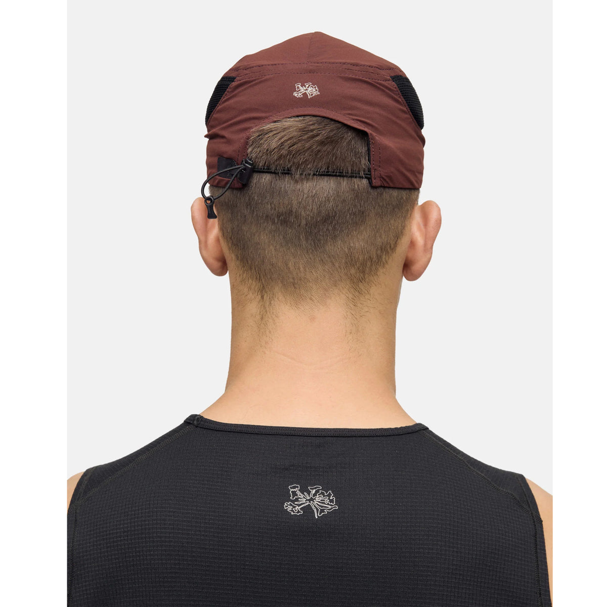 A person is seen from behind, wearing a dark sleeveless top and the Portal Planar Lightweight Cap in Bitter Chocolate. Both the top and cap feature a white embroidered Portal logo, and the cap includes a small adjustable cord for a customizable fit. The background is plain and light-colored.