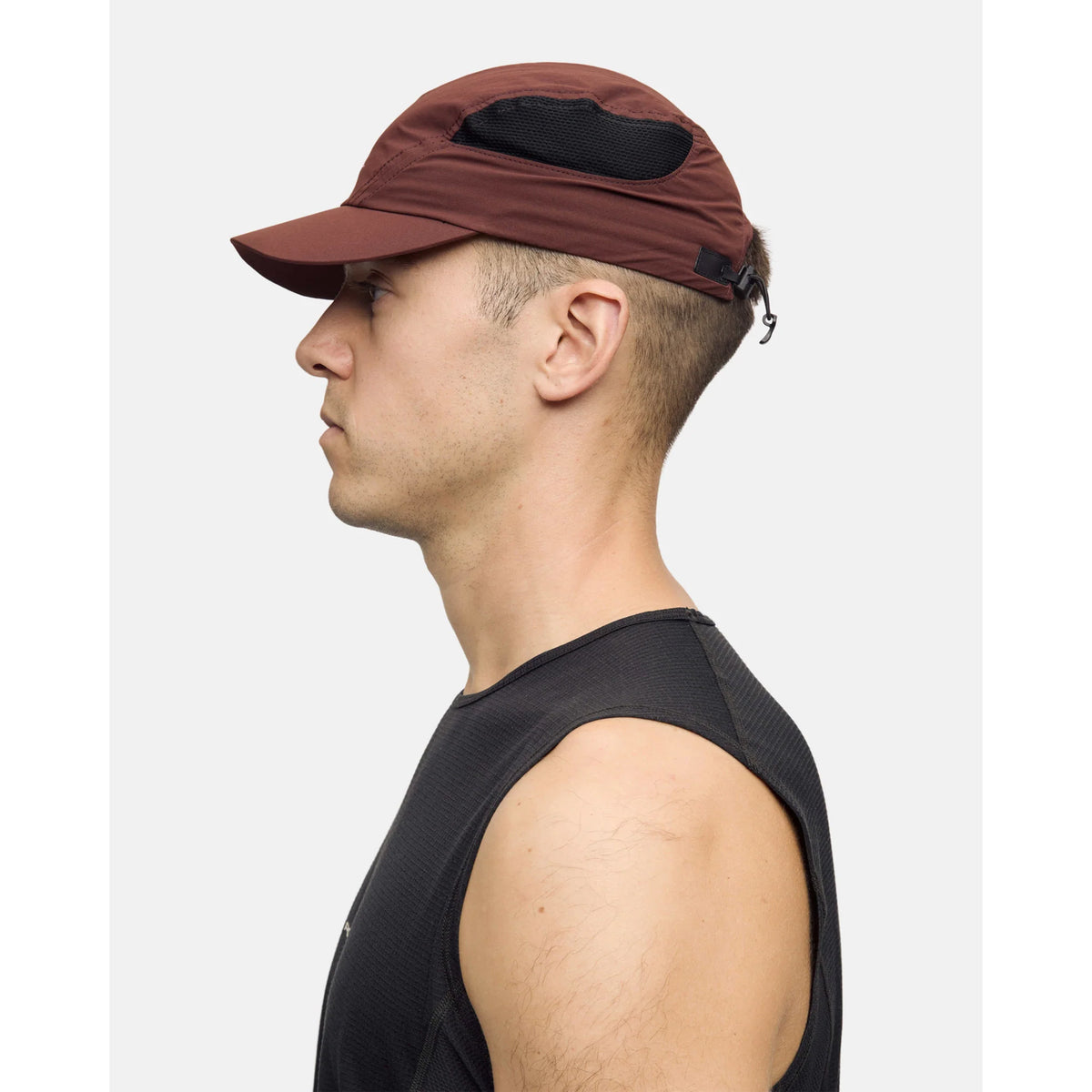 A man wearing the Portal Planar Lightweight Cap in Bitter Chocolate, featuring black mesh detailing, and a black sleeveless athletic shirt, viewed in profile against a plain light background.