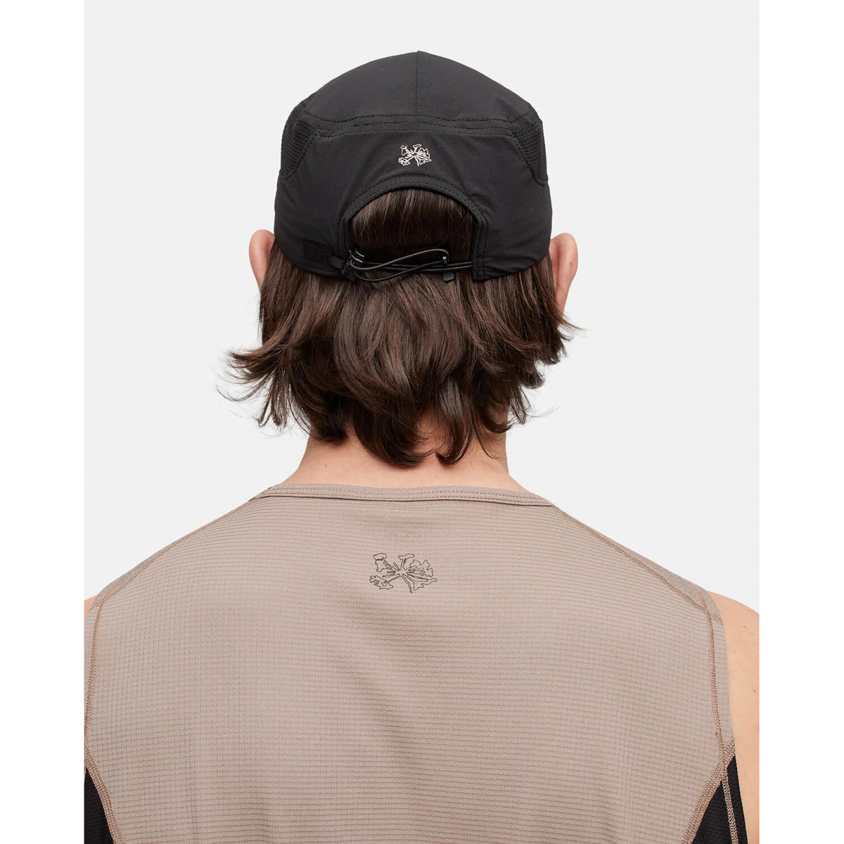 A person with medium-length brown hair is shown from the back against a plain background, wearing the Portal Planar Lightweight Cap in Black Beauty—a breathable black cap with an adjustable fit and a small embroidered logo—paired with a light brown textured sleeveless shirt that also features subtle embroidered branding.