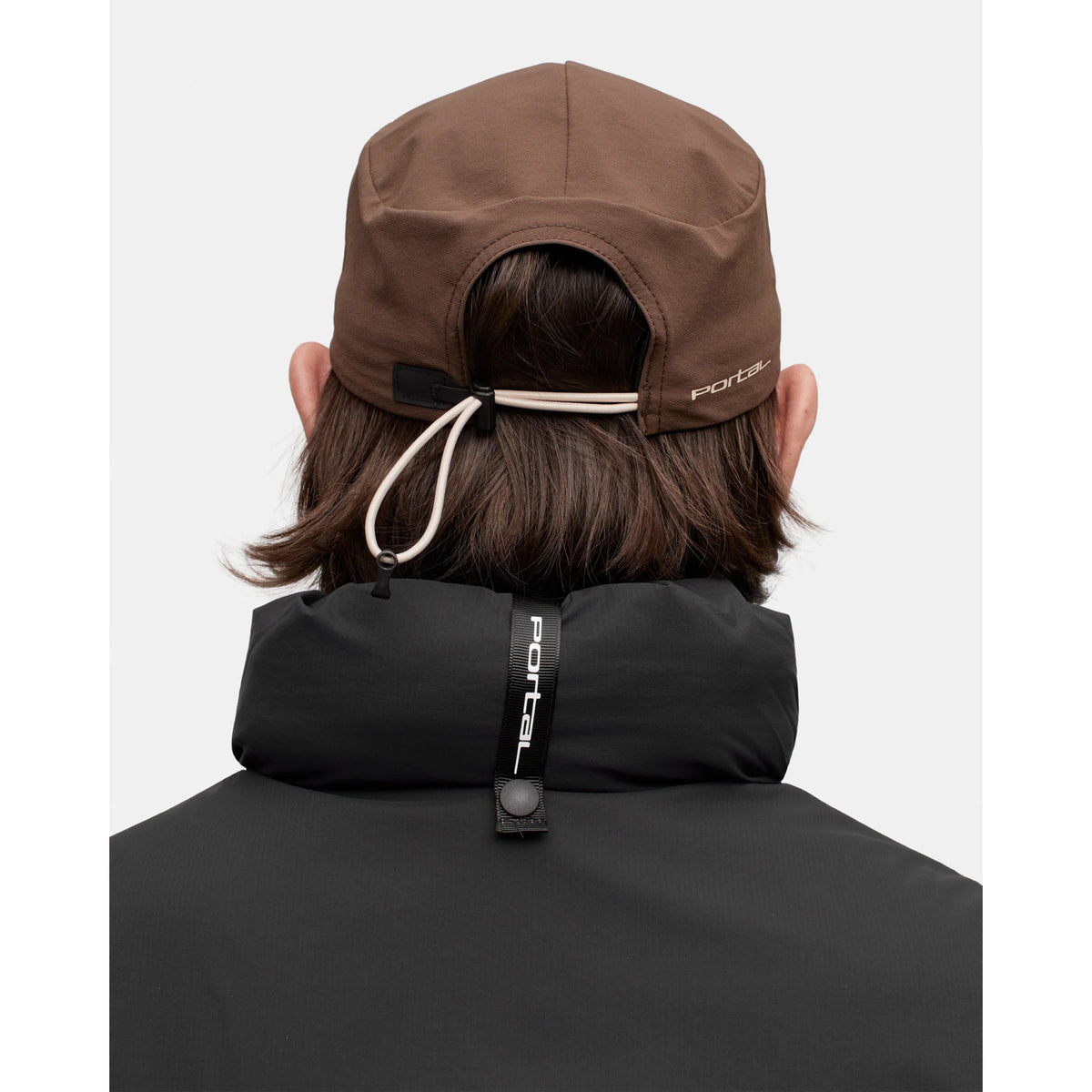 A person with shoulder-length brown hair is seen from behind, wearing the Portal Radius Cap in Slate Black—a lightweight, active-use cap made from durable double weave nylon stretch fabric for comfort—along with a black Portal jacket. The cap features a drawstring adjustment and both items display the Portal logo, visible on the back of the head and upper back.