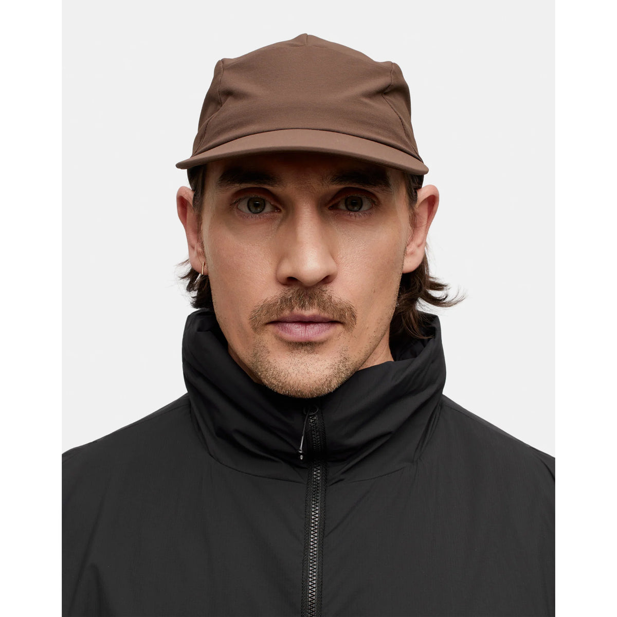A person with light skin, brown hair, and a mustache looks directly at the camera against a plain white background, wearing the Portal Radius Cap in Slate Black—an active use cap crafted from double weave nylon stretch material—paired with a black zip-up jacket.