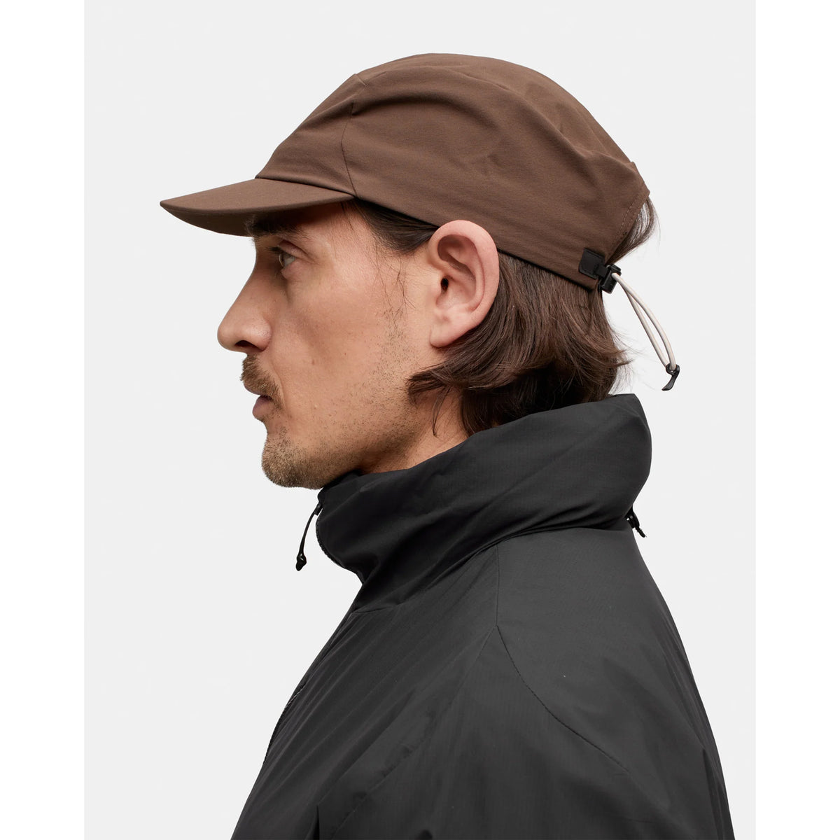 A man in profile wears the Portal Radius Cap in Slate Black, featuring a lightweight design with a drawstring at the back, along with a black high-collar jacket, set against a plain light background.