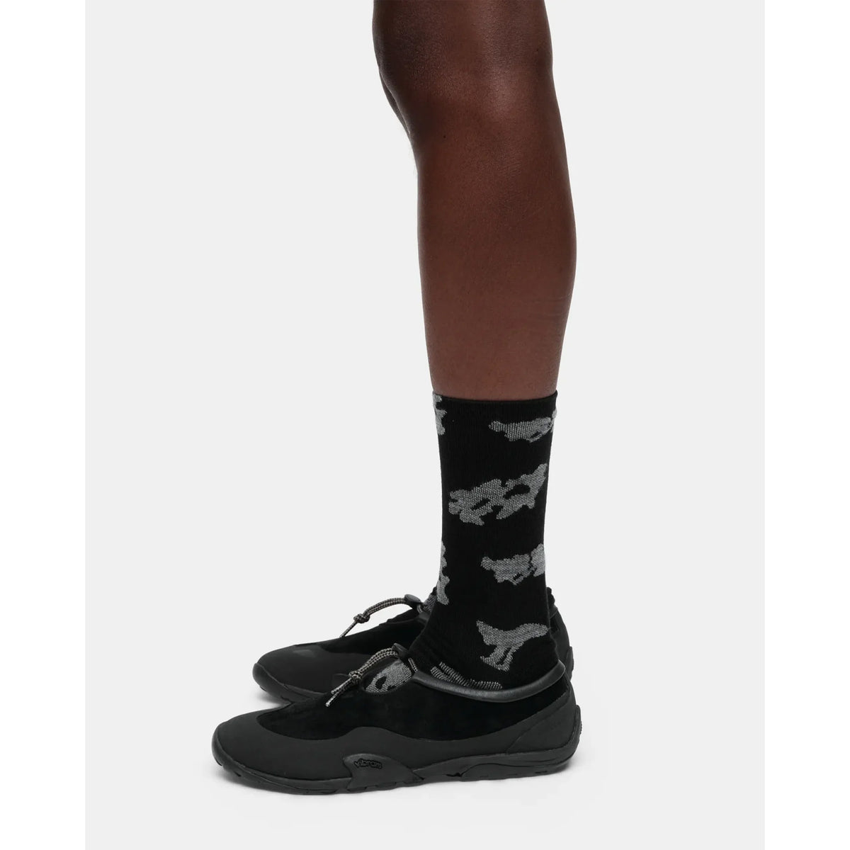 A person wearing black shoes with toggle laces and Portal Ritual Merino Socks Black Beauty, featuring a gray animal pattern, stands against a plain white background.