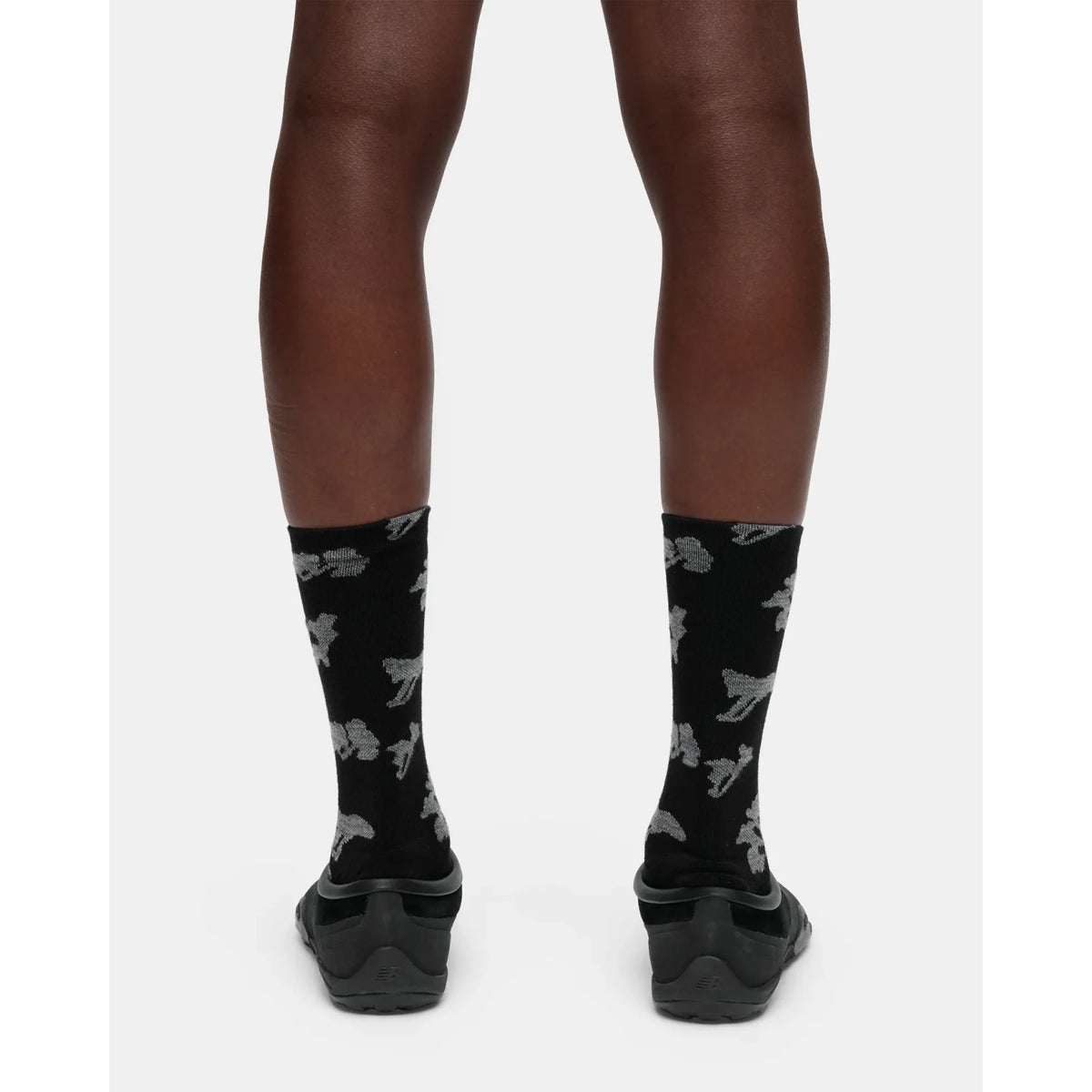 A person wearing Portal Ritual Merino Socks in Black Beauty, featuring a gray abstract pattern, and black slip-on shoes, shown from the back against a plain white background.