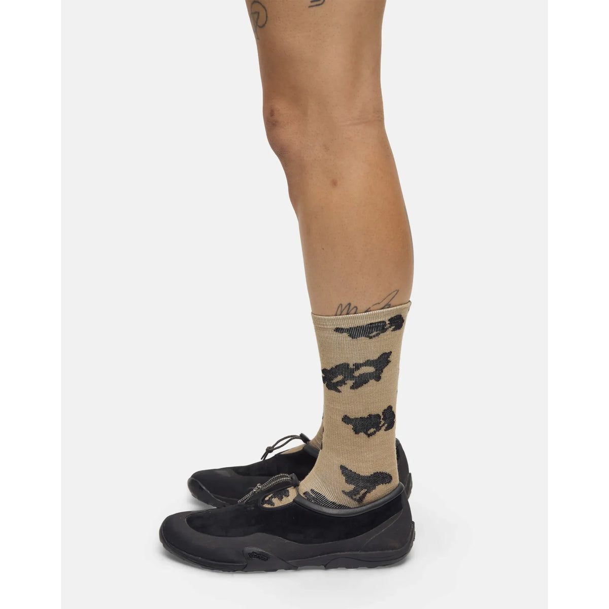 A person stands sideways against a white background, wearing Portal Ritual Merino Socks in Greige featuring black cloud patterns, paired with black shoes. Tattoos are visible on their legs and ankles.