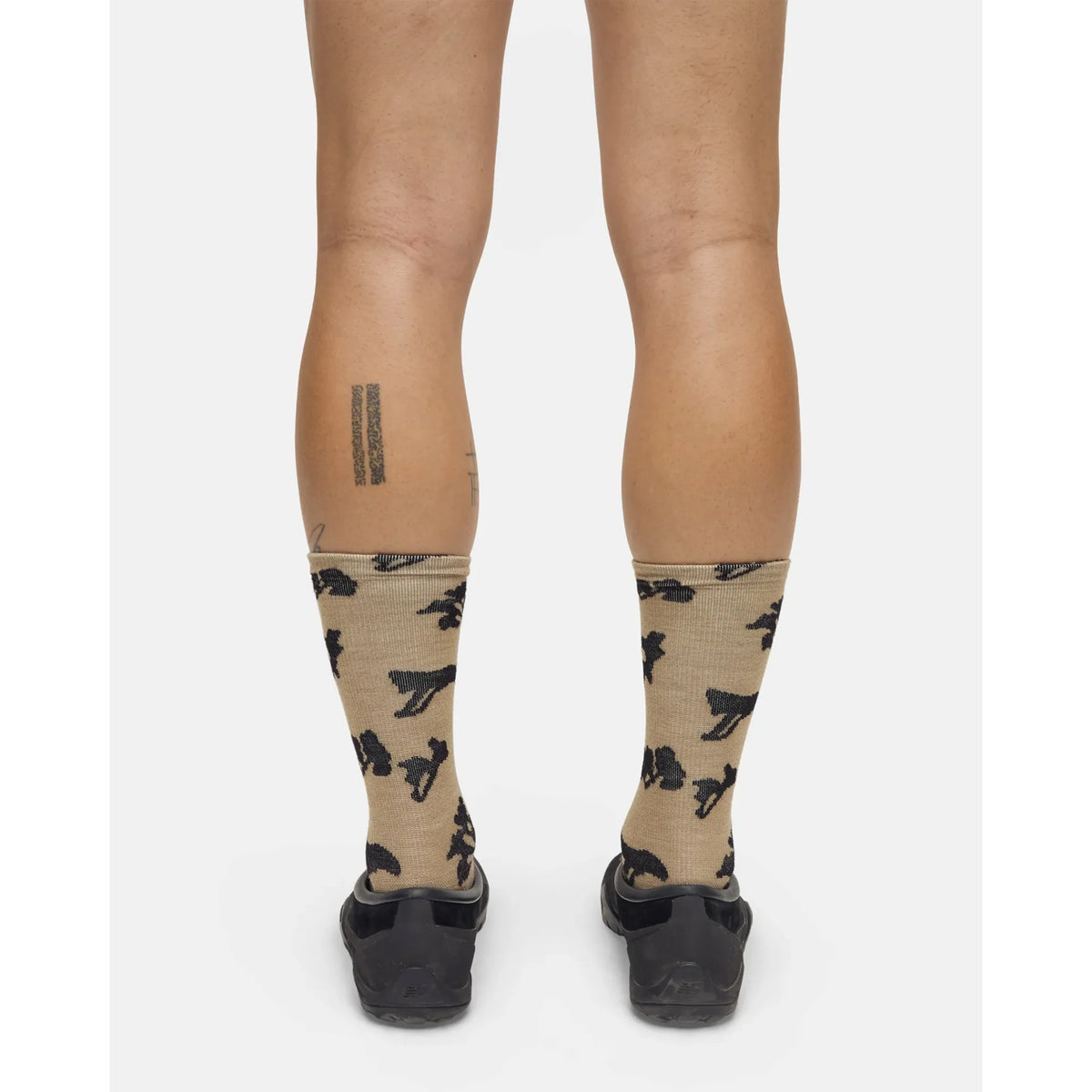 A person stands with their back to the camera, wearing Portal Ritual Merino Socks in Greige featuring black patterns and paired with black shoes. A rectangular tattoo is visible on their left calf, while a faint tattoo appears on their right calf.