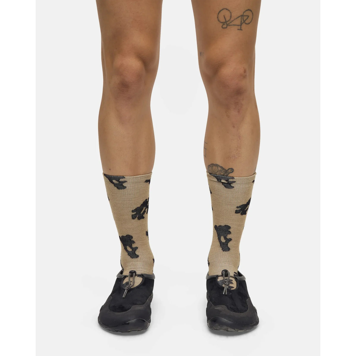 A person wears Portal Ritual Merino Socks in Greige, featuring durable construction and black bear designs, paired with black athletic shoes and showing a bicycle tattoo on one knee. The photo highlights their legs against a plain white background.