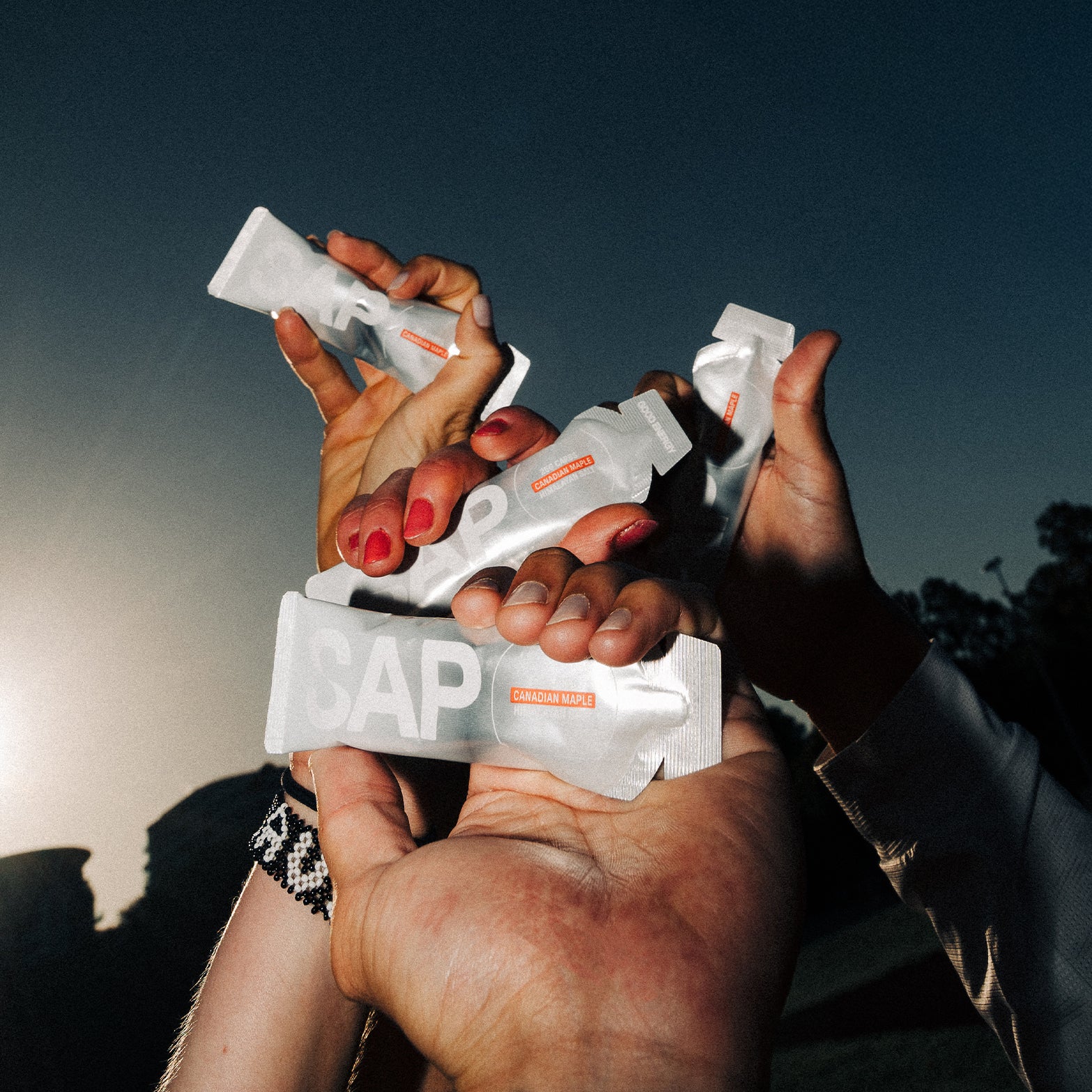 Four hands hold up white packages labeled SAP against a backdrop of dark sky and sunset, with one hand wearing a black and white bracelet—each SAP25 Energy Gel from the box of 10 delivers pure Canadian maple syrup fuel in an energy gel with a 50:50 carb ratio for balanced performance.