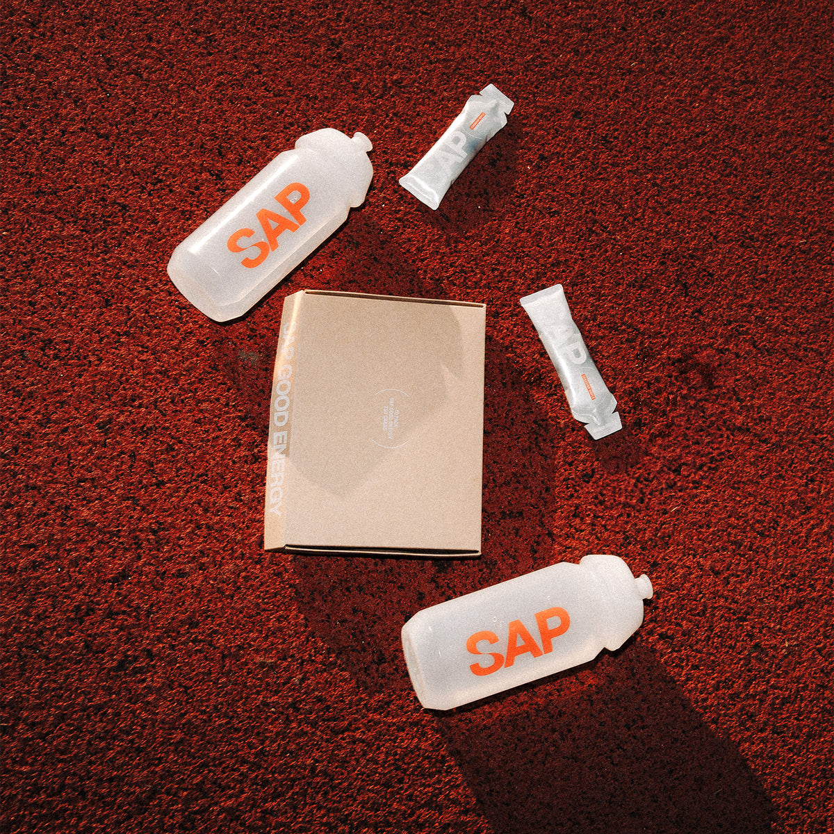 Two clear water bottles labeled SAP, a brown box, and three SAP energy gel packets are placed on a textured red surface in bright sunlight.