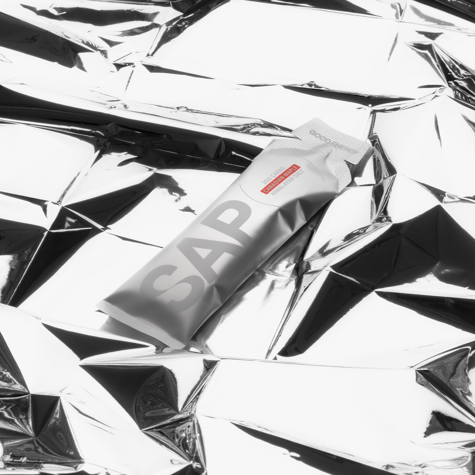 A silver SAP25 Energy Gel box by SAP rests on crumpled, reflective silver foil, creating a modern, shiny, and minimalist look—emphasizing its maple syrup fuel and balanced 50:50 carbohydrate ratio for sustained energy.