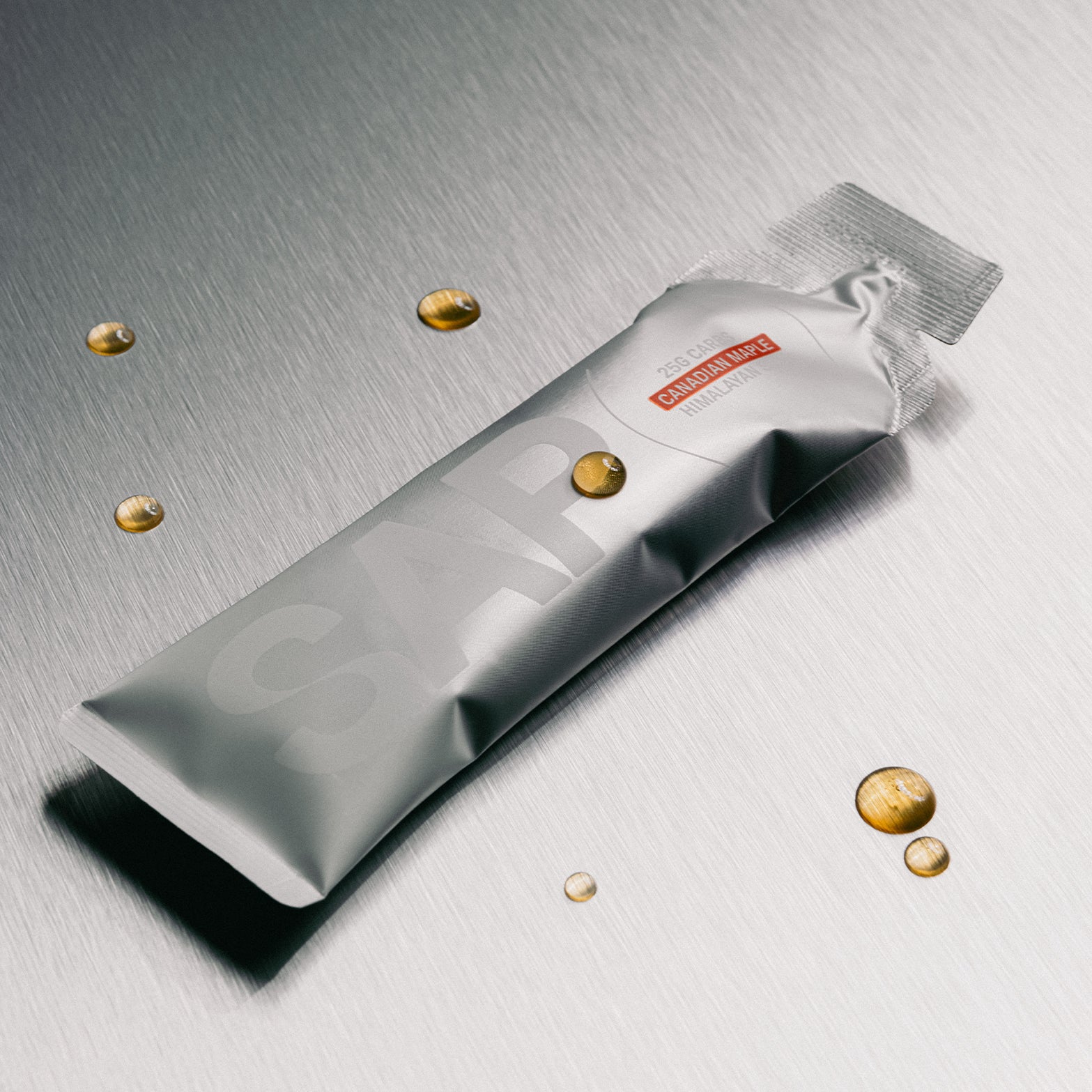 A silver sachet from SAP labeled “SAP25 Energy Gel” rests on a metallic surface, surrounded by small droplets of golden liquid that evoke its maple syrup fuel and balanced 50:50 carb ratio—an energy gel expertly crafted by SAP for rapid replenishment, available in a box of 10.