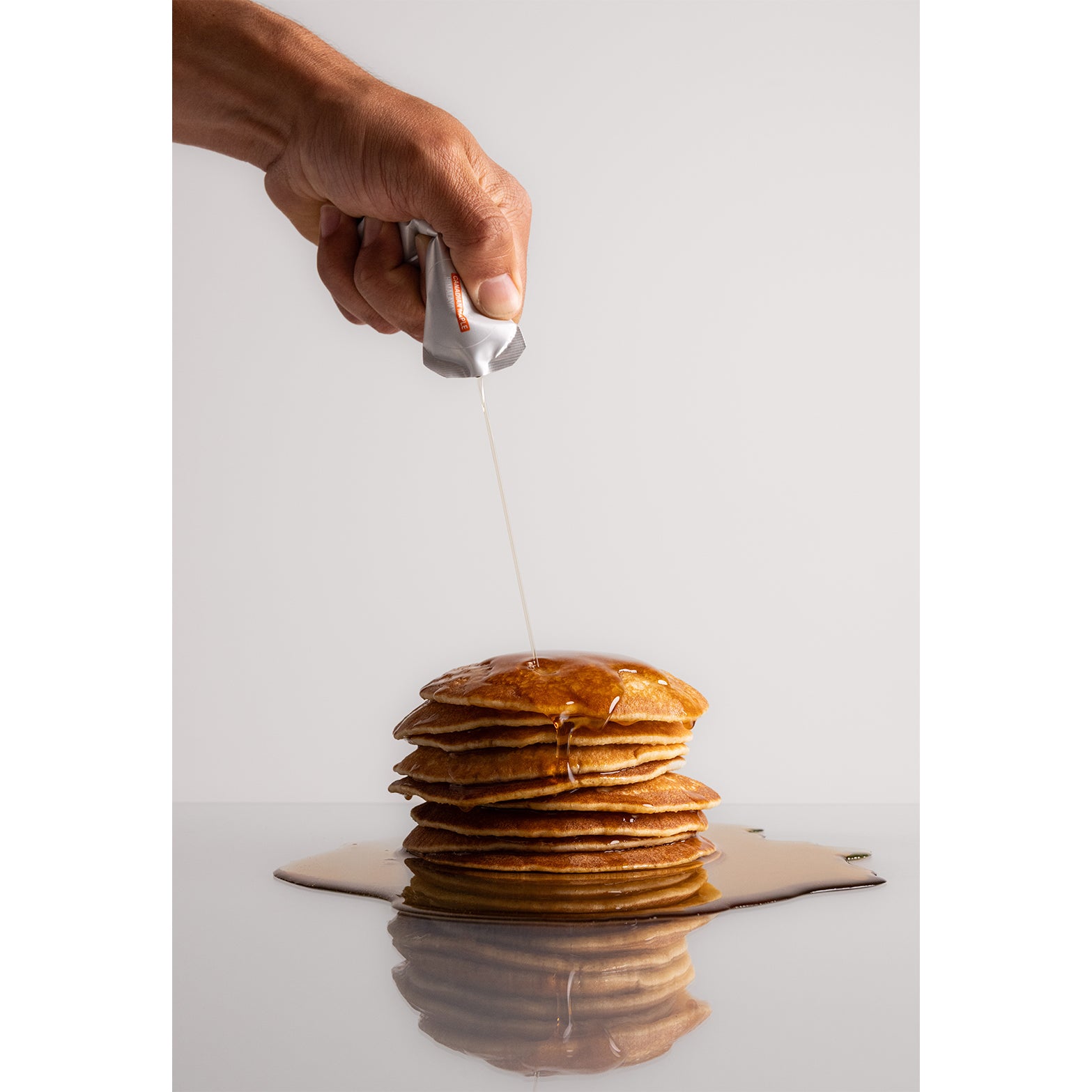 A hand pours SAP25 Energy Gel from a packet onto a stack of pancakes, the energy gel pooling around the base on a reflective surface—reminiscent of maple syrup fuel with a balanced 50:50 carb ratio from SAP, delivering that extra boost for your morning.