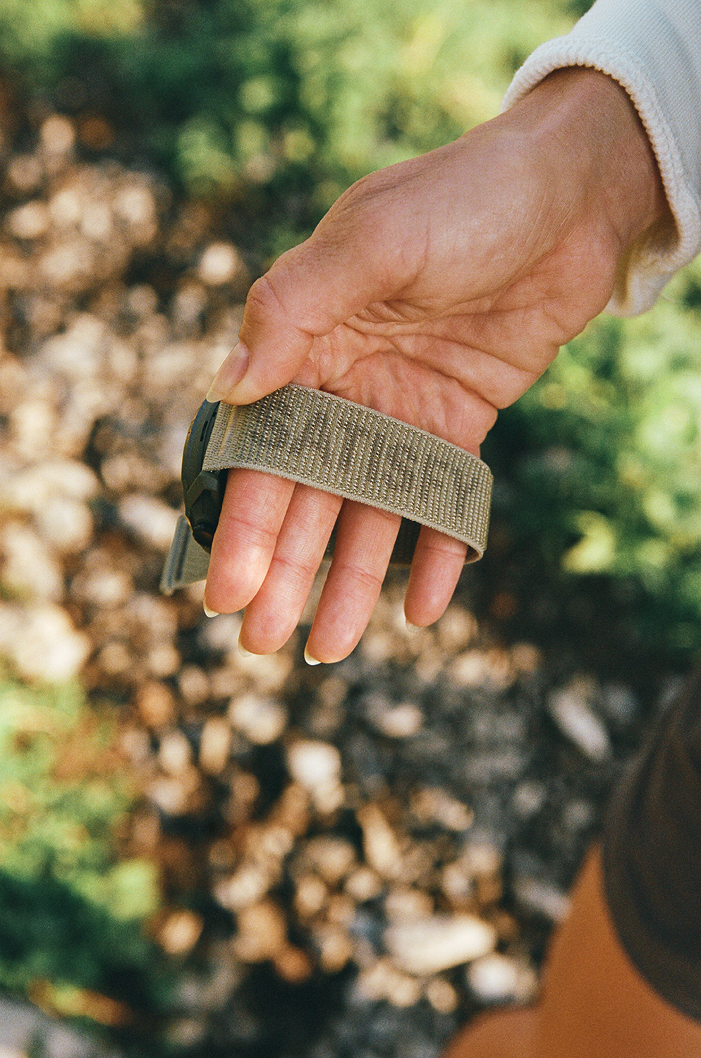 A hand with neatly trimmed nails holds a khaki-colored fabric watch strap outdoors, with blurred greenery and stones in the background.