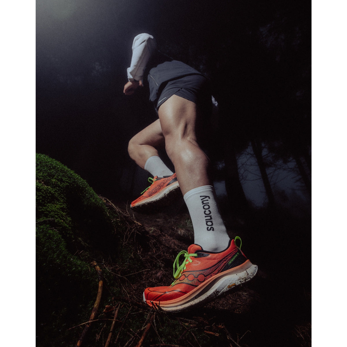 A runner in bright orange trail shoes and white socks with SAUCONY text climbs a mossy, forested hill at night, seen from a low angle.