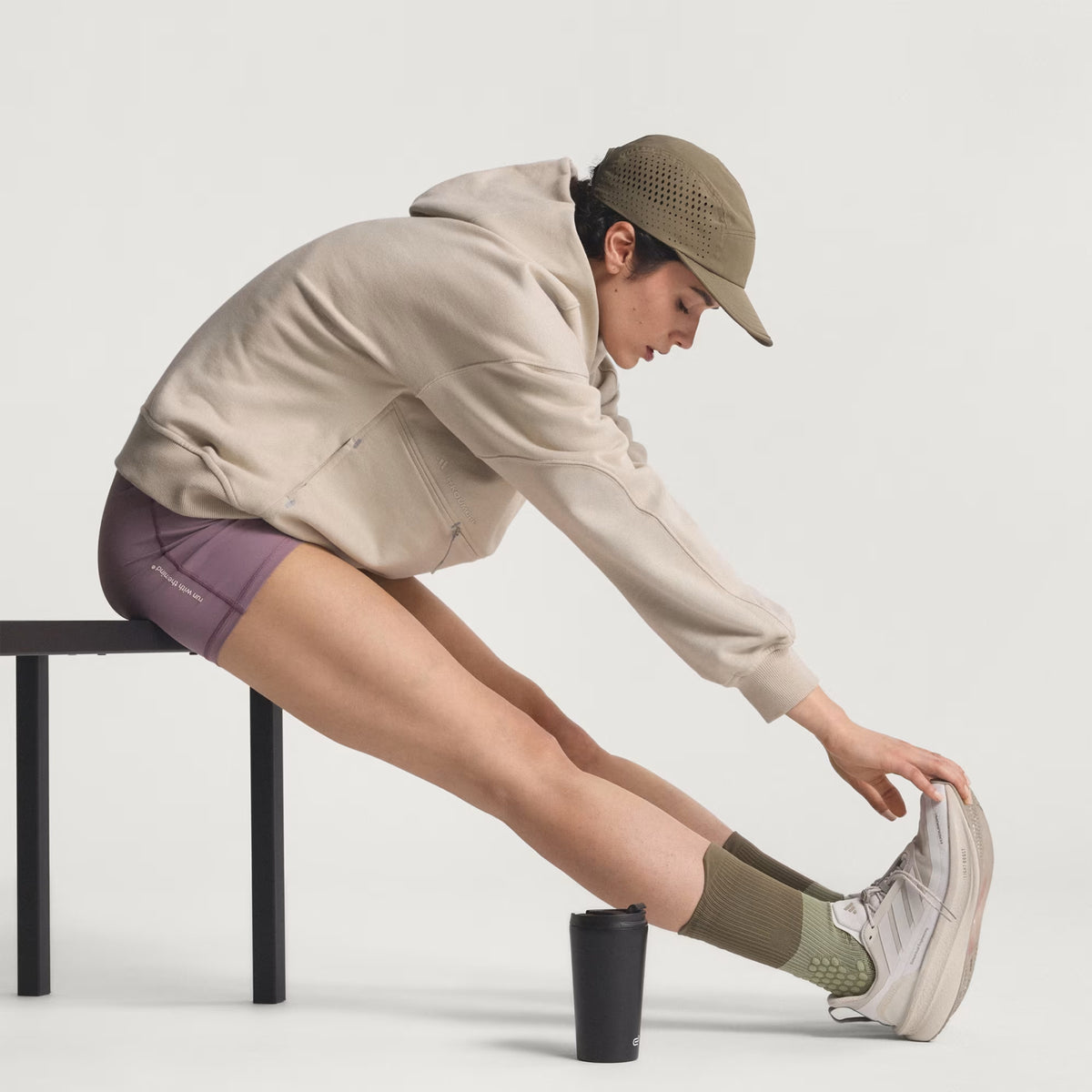 Person in a beige hoodie, purple shorts, cap, and green socks stretches one leg on a bench with a black tumbler next to them, against a plain white background.