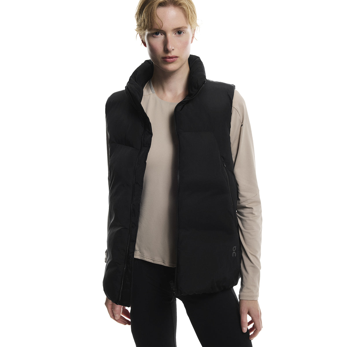 A person with short blond hair wears a beige long-sleeve shirt and a black puffer vest while standing against a plain white background.