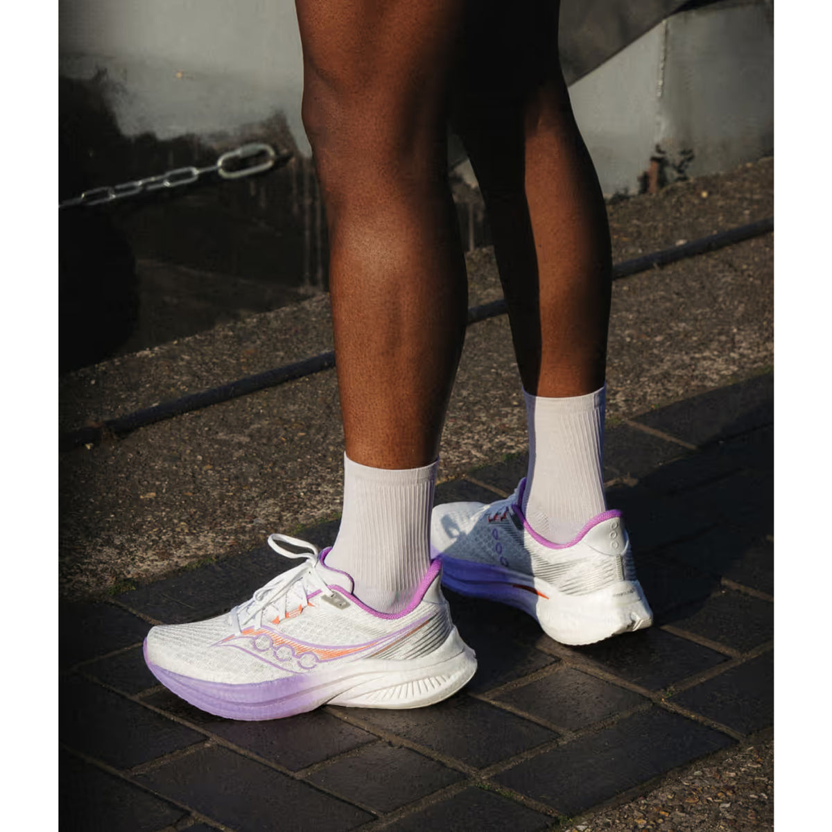 Person standing outdoors on a paved surface, wearing white running shoes with purple accents and white crew socks. Only their legs from knees down are visible.