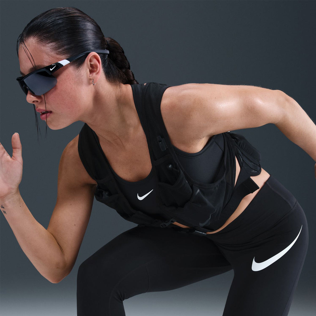 A woman in black athletic wear and sunglasses is in a running pose against a dark background. She is wearing a Nike sports bra, leggings, and a weighted vest.
