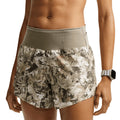 A person wearing Nike Women’s ACG Chamo Camo Dri-FIT 10cm Brief-Lined Running Shorts in Sequoia / Summit White and a watch, with their hands resting by their sides. The background is plain white.