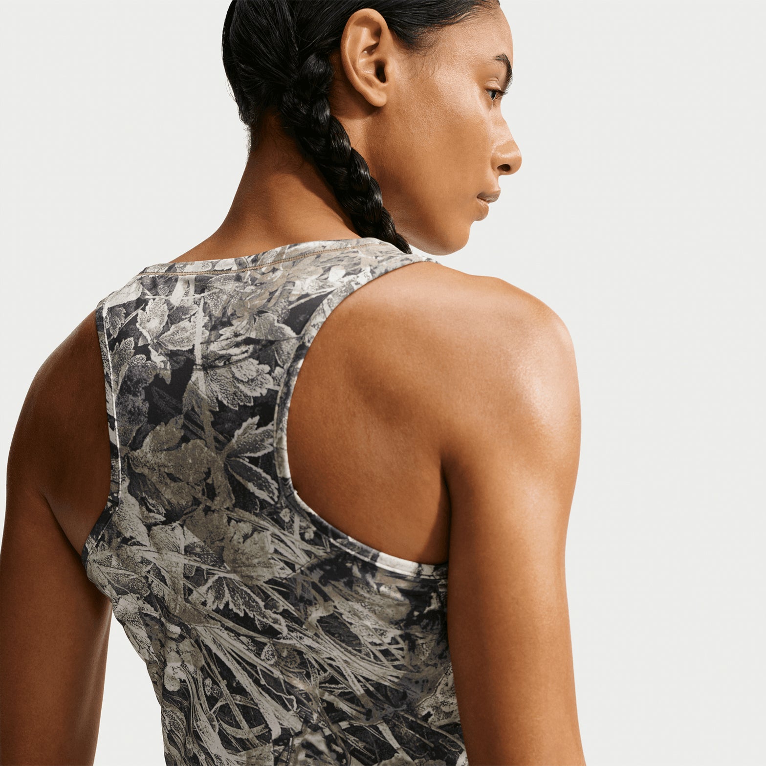 A person with braided hair is shown from the back, wearing a Nike Women’s ACG Chamo Camo Dri-FIT Tank Top in Sequoia and Summit White. The background is plain and light-colored.