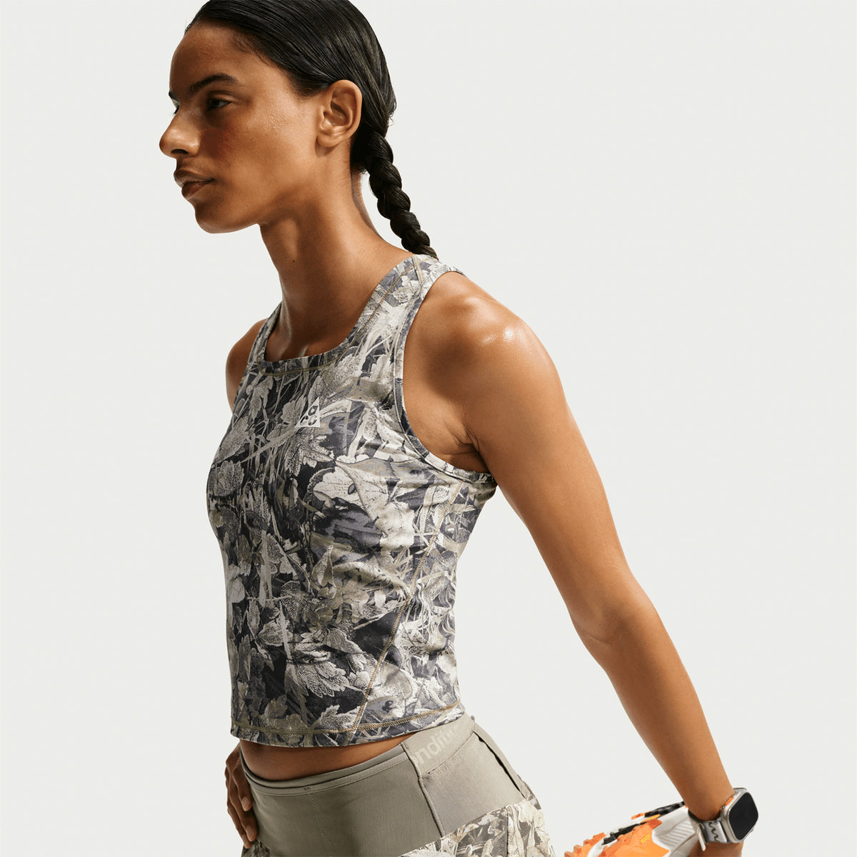 A person with a braided hairstyle stretches their arms back, wearing a sleeveless, patterned athletic top and gray workout pants against a plain background.
