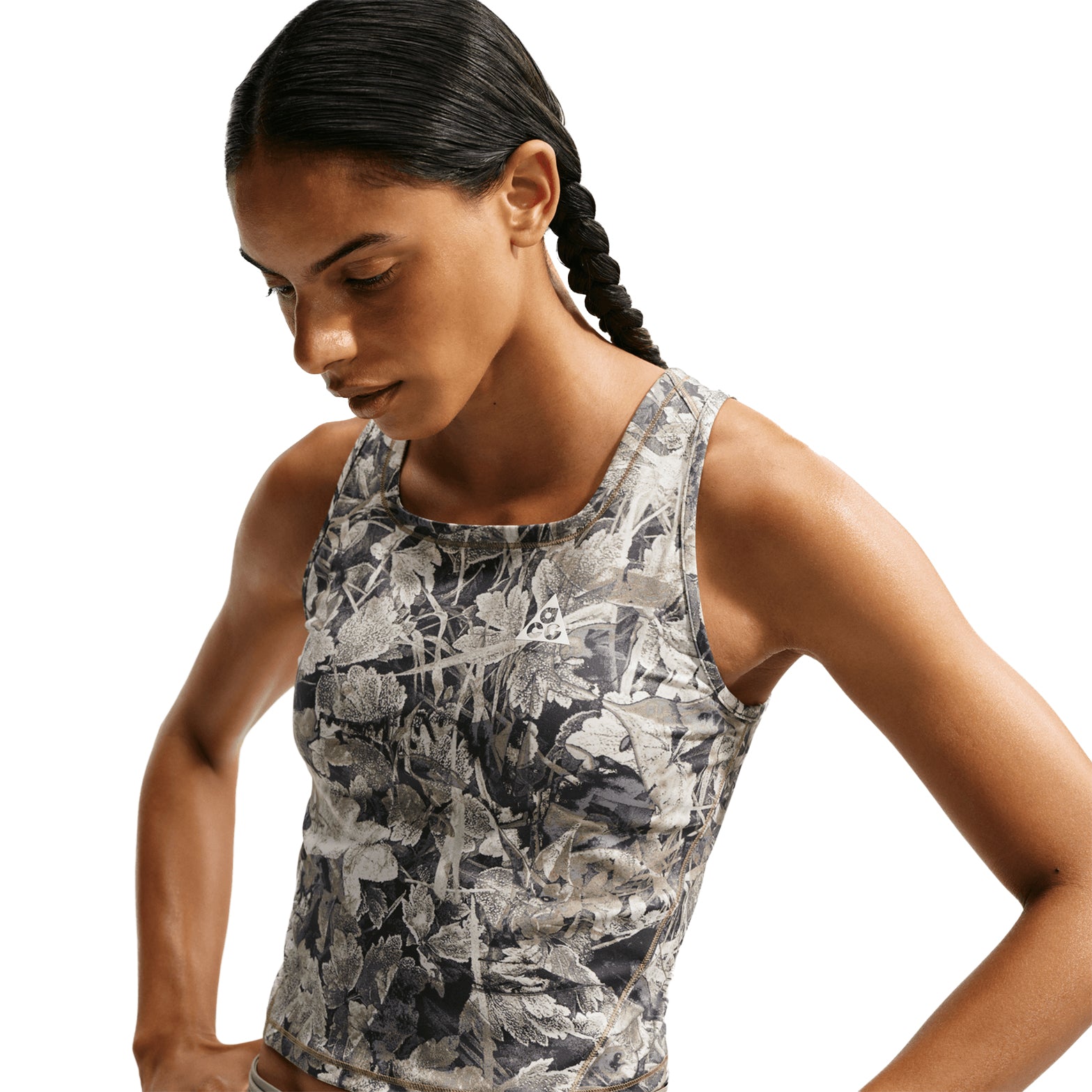 A person with dark braided hair wears the Nike Women’s ACG Chamo Camo Dri-FIT Tank Top in Sequoia/Summit White, featuring a camo-inspired leafy pattern, while looking down thoughtfully against a plain white background.