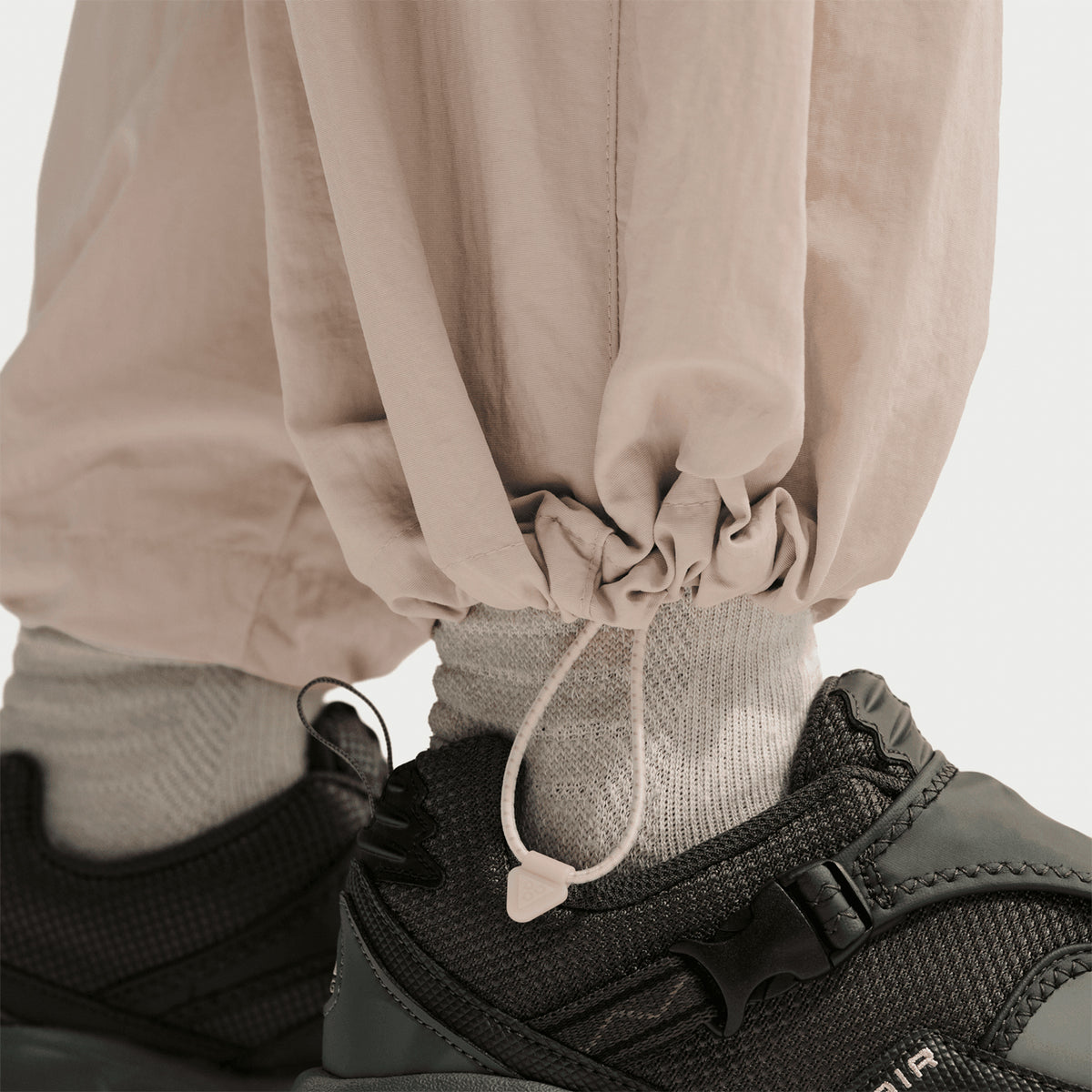 Close-up of a person wearing light beige drawstring pants, grey socks, and dark athletic sneakers. The drawstring at the pant cuff is cinched, and the background is plain and light-colored.