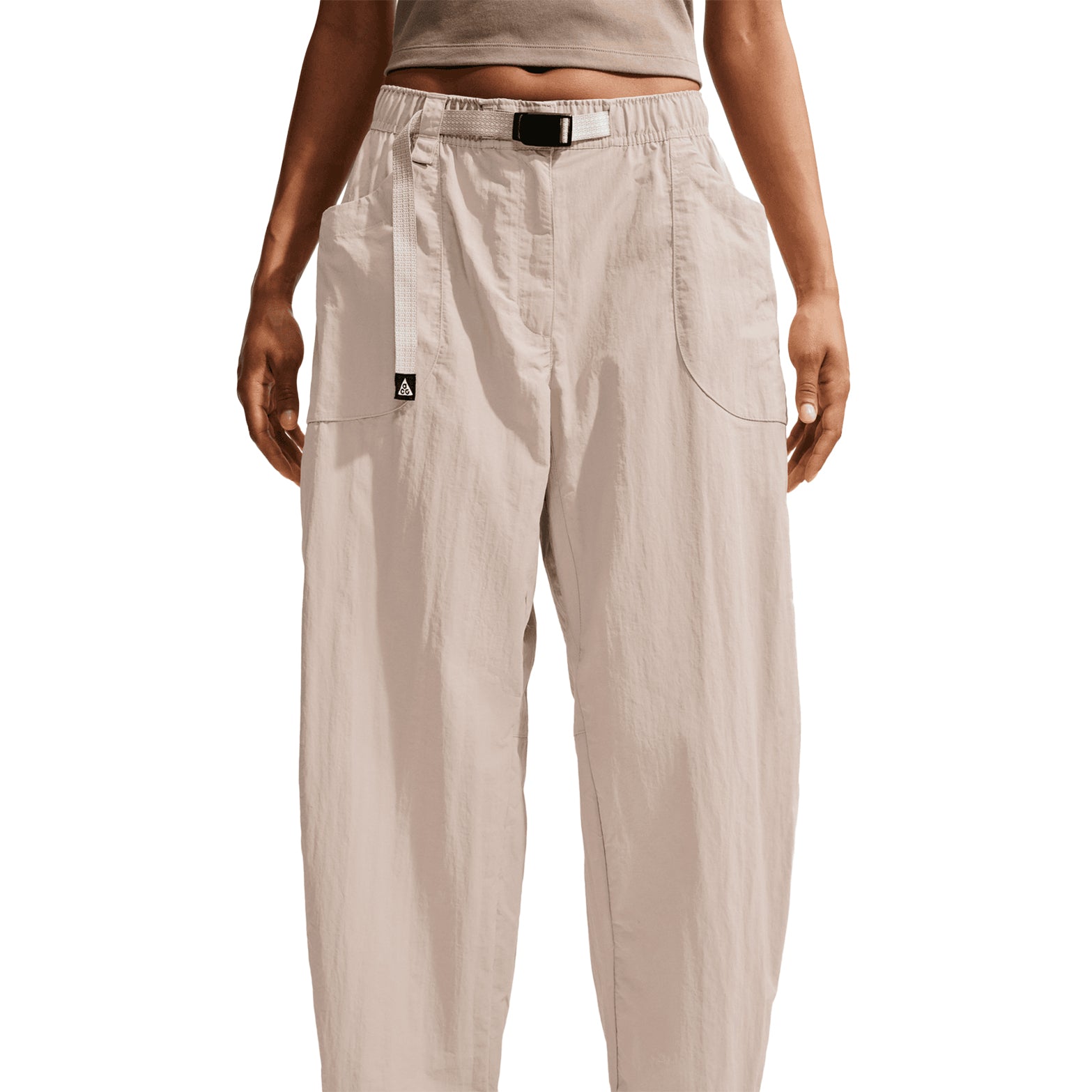 A person is shown from mid-torso to feet against a white background, wearing Nike Women’s ACG Dolomiti Trousers in Cream II / Light Orewood Brown / Summit White. The high-waisted, loose-fitting mountain hiking trousers feature large front pockets and a black belt.