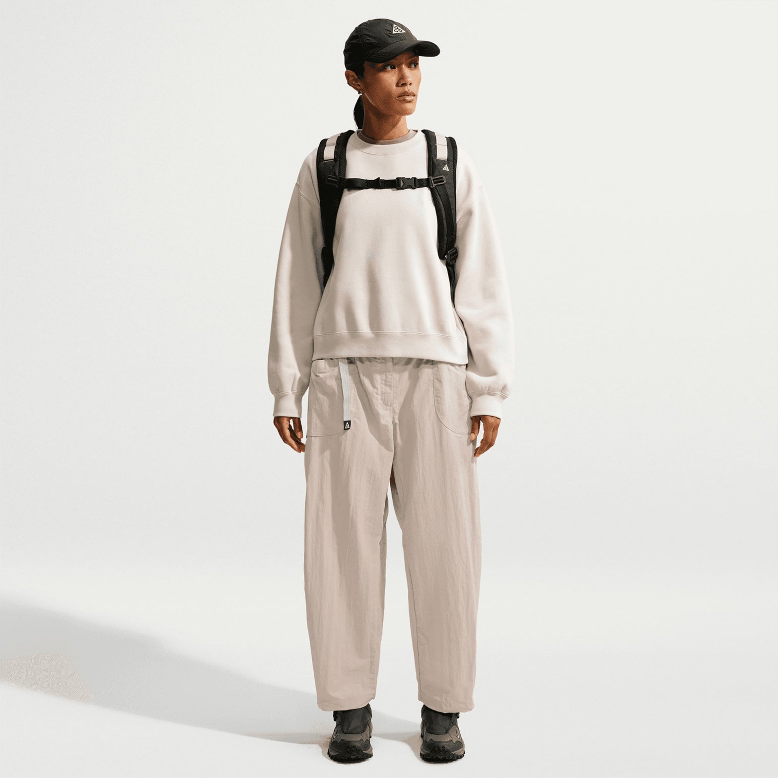A person wearing a black cap, white sweatshirt, Nike Women’s ACG Dolomiti Trousers in Cream II / Light Orewood Brown / Summit White, black shoes, and a black backpack stands against a plain white background.