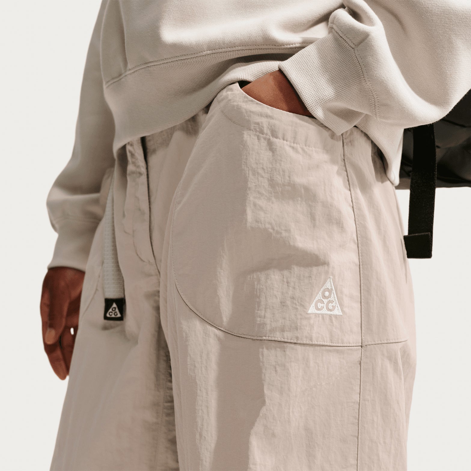 A person wearing Nike Women’s ACG Dolomiti Trousers in Cream II/Light Orewood Brown/Summit White and a light sweatshirt stands with one hand in their pocket, while a black bag strap is visible over their shoulder.