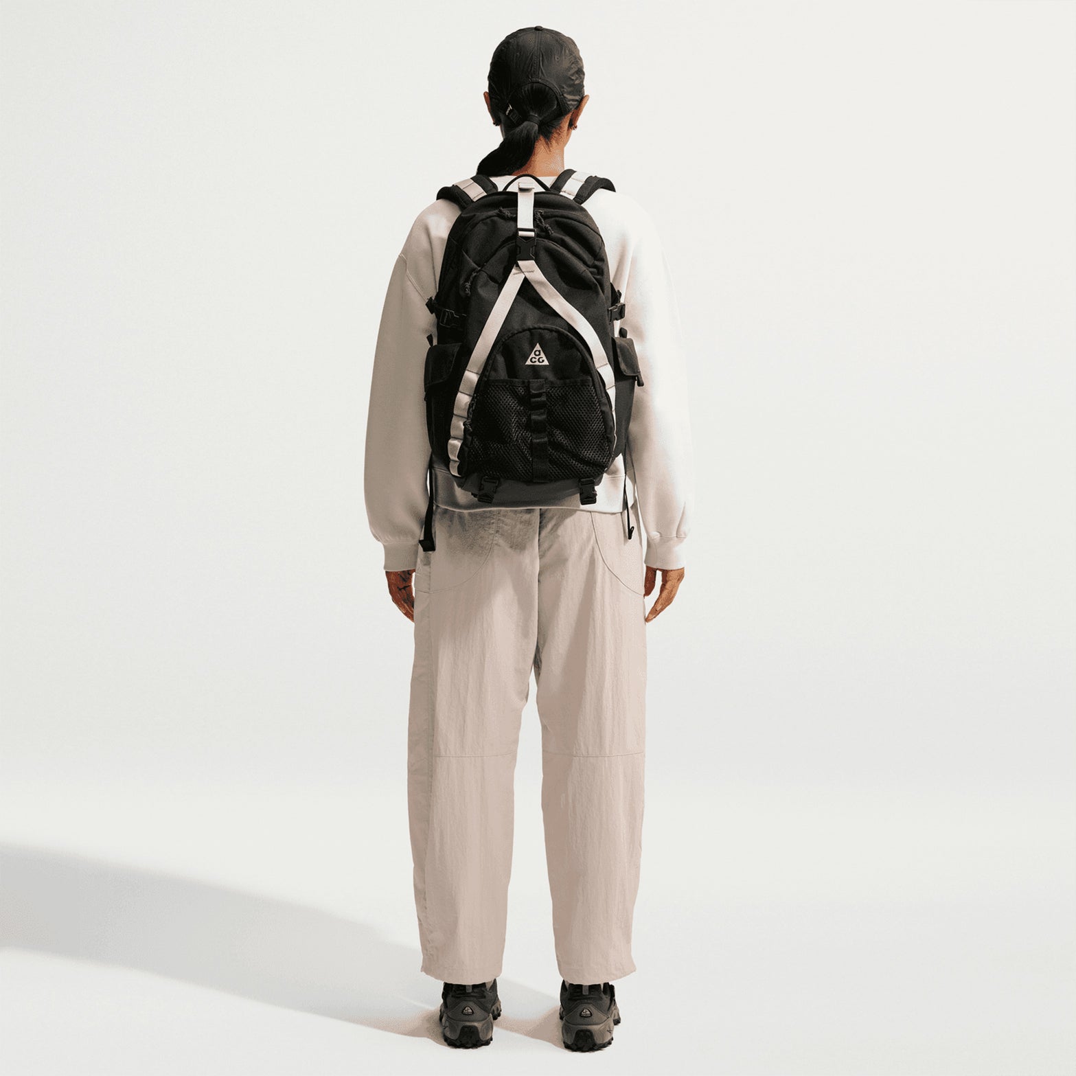 A person stands facing away, wearing a black backpack, a white long-sleeve shirt, black shoes, and Nike Women’s ACG Dolomiti Trousers in Cream II / Light Orewood Brown / Summit White against a plain white background.