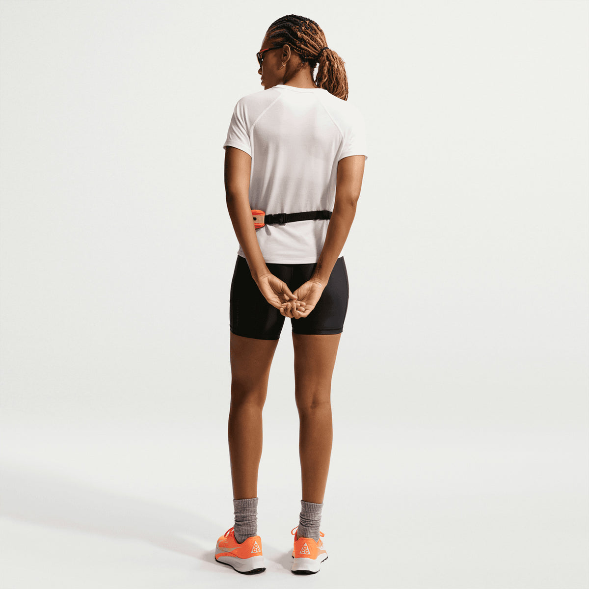 A woman with braided hair stands facing away, wearing the Nike Women’s ACG Dri-FIT Trail Running Top in Phantom / Summit White, along with black shorts, gray socks, and bright orange sneakers. Her hands are clasped behind her back and she wears sunglasses.