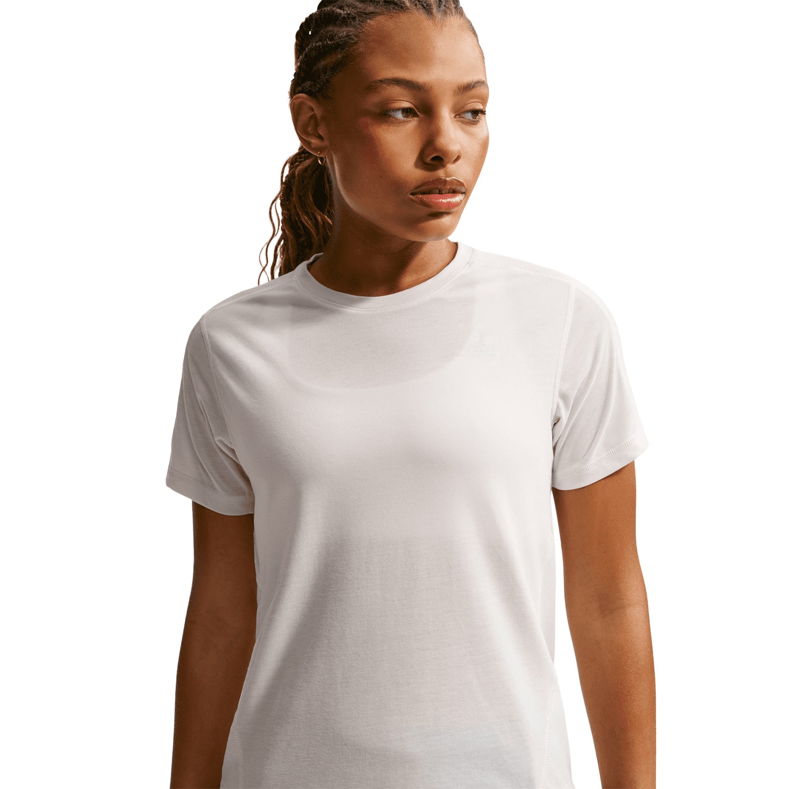 A woman with braided hair stands against a white background, looking to the side with a neutral expression, wearing the Nike Women’s ACG Dri-FIT Trail Running Top in Phantom/Summit White. The top features Dri-FIT technology for enhanced comfort during outdoor activities.