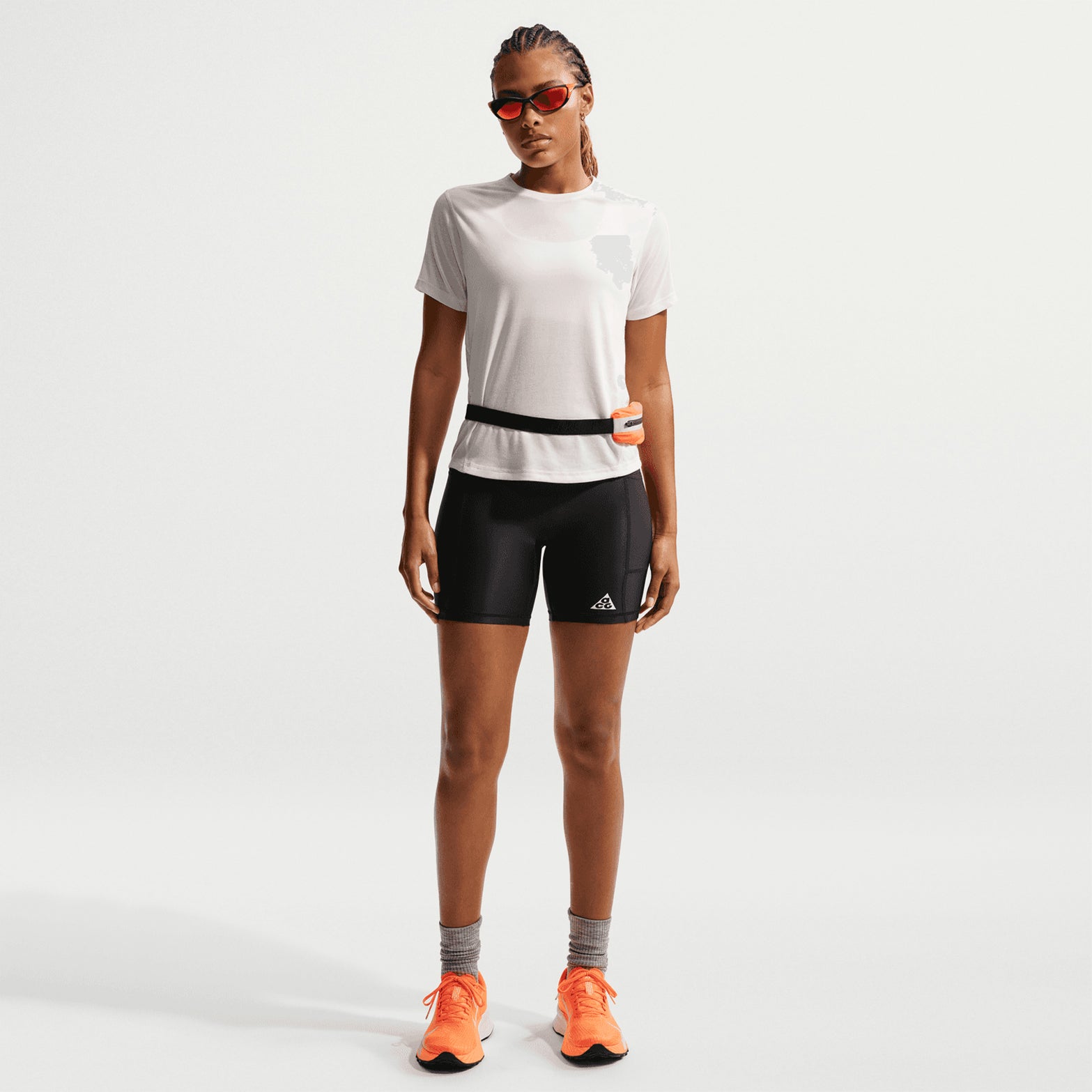 A person is standing facing forward, wearing the Nike Women’s ACG Dri-FIT Trail Running Top in Phantom / Summit White, paired with black shorts, gray socks, bright orange running shoes, red sunglasses, and a waist belt.