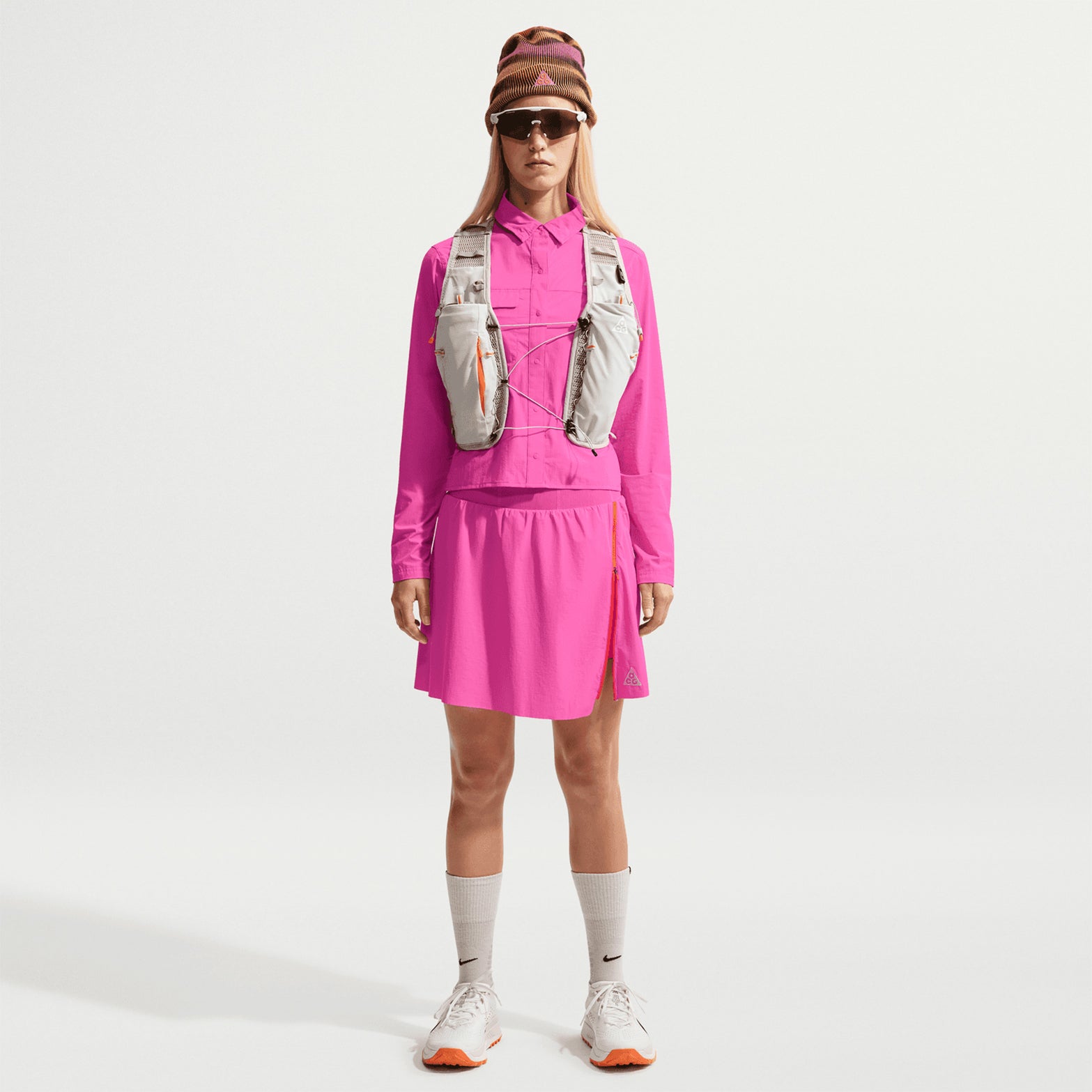 Women's Nike ACG High Stakes Skort Desert Pink Light Crimson Summit White front full body view on female trail runner showing stretch-woven outer with built-in knit shorts, UV protection and zippered design for hiking and trail activities