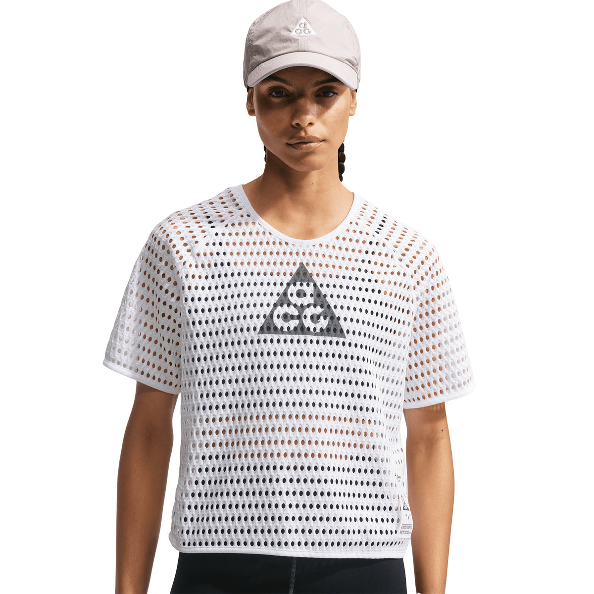A woman wears the Nike Women’s ACG Radical AirFlow Tee White / Anthracite, featuring a triangular logo on the front and a light-colored cap, against a plain white background.