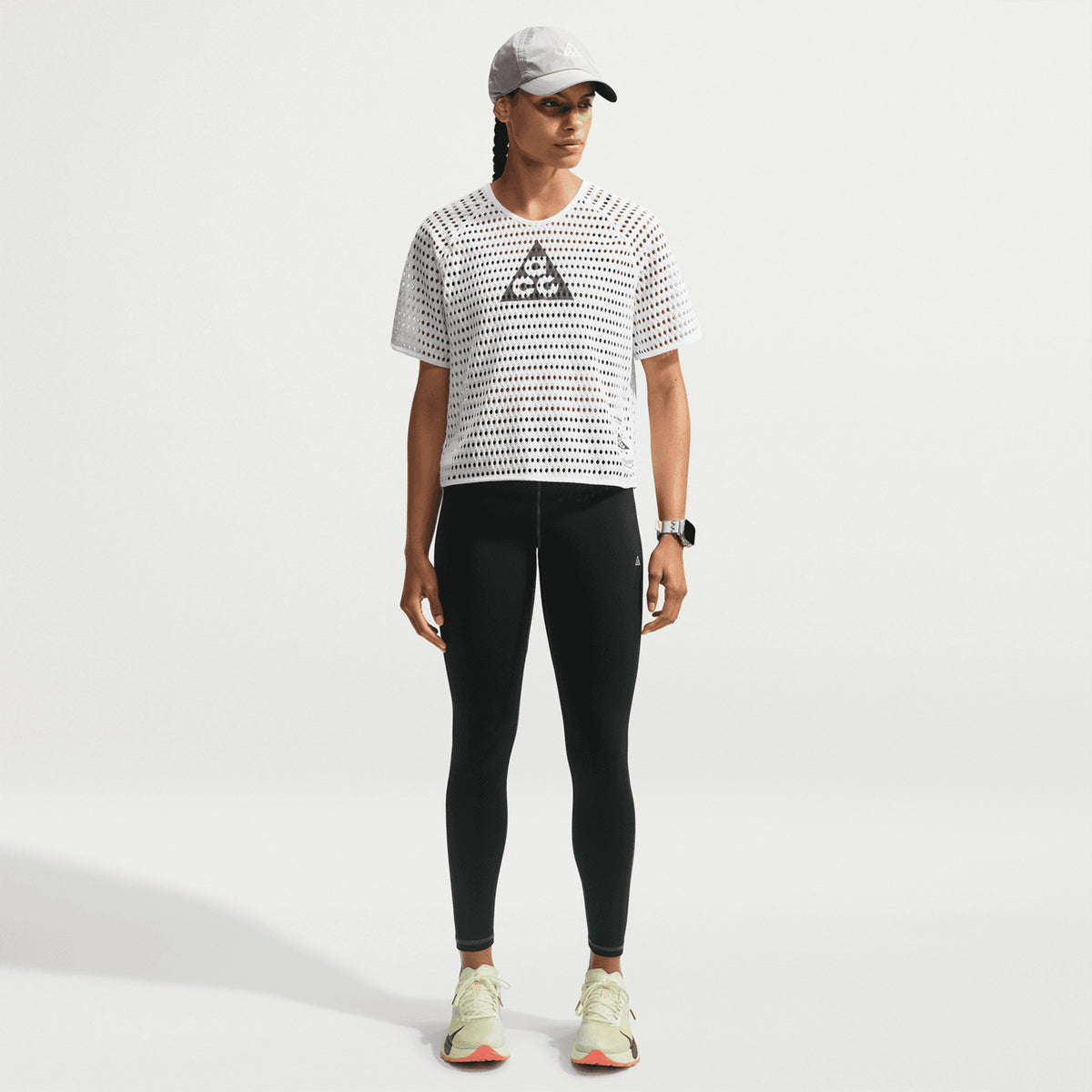 A person in a gray cap, Nike Women’s ACG Radical AirFlow Tee White / Anthracite, black leggings, and light sneakers stands against a plain white background.