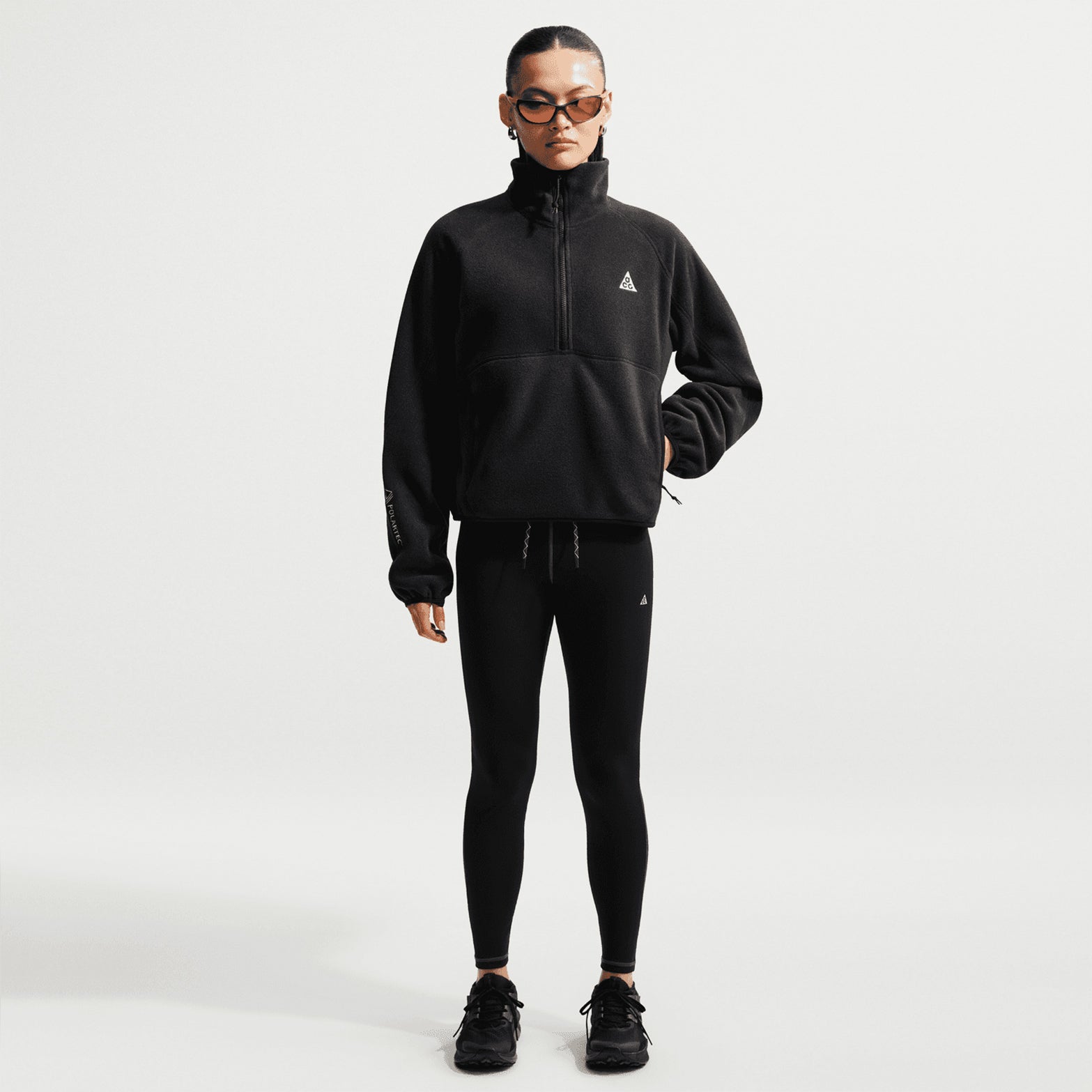 A person wearing black sunglasses, black leggings, and black sneakers stands against a plain white background, styled in the Nike Women’s ACG Wolf Tree 1/2 Zip Top in Black / Anthracite / Summit White.