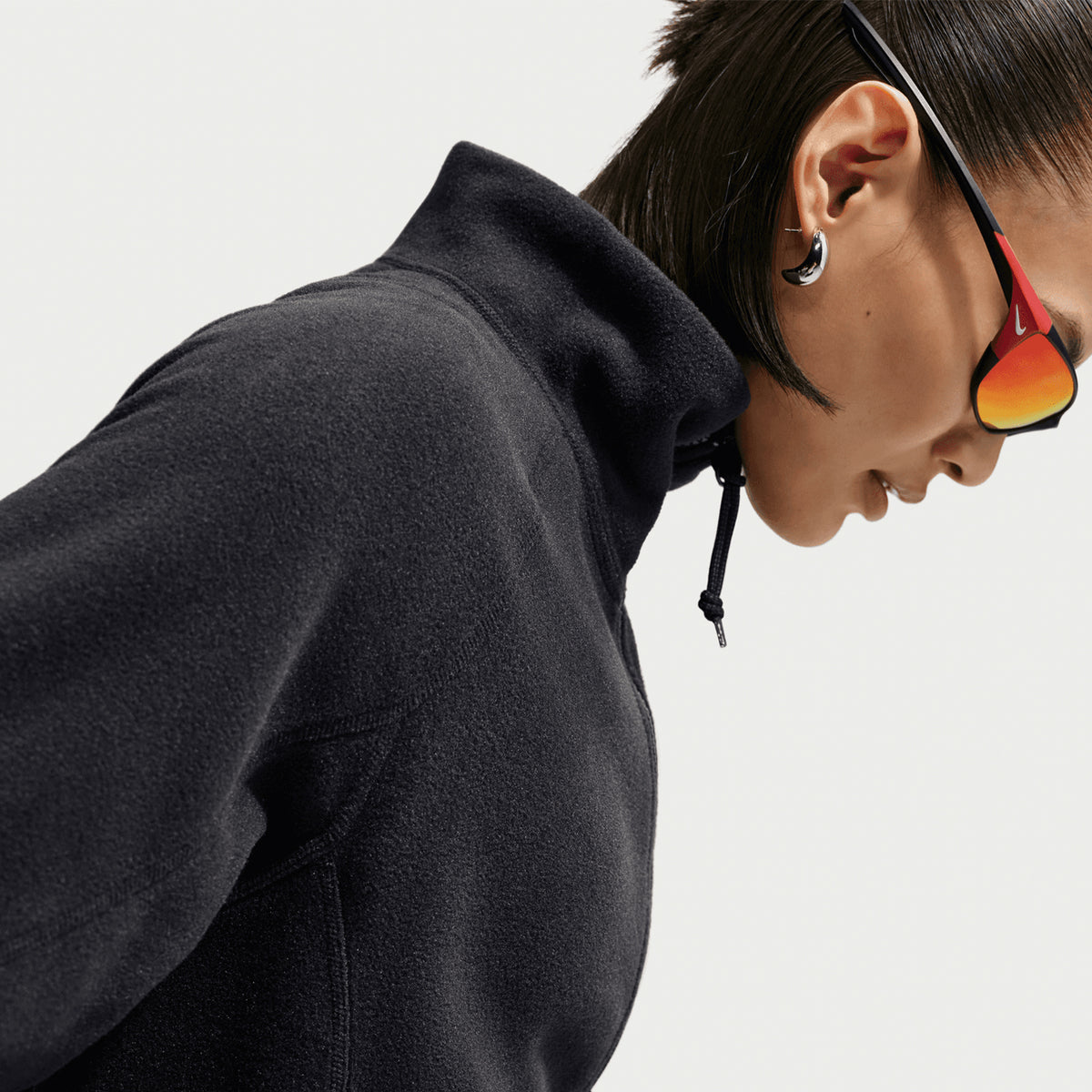 A person wearing the Nike Women’s ACG Wolf Tree 1/2 Zip Top in Black / Anthracite / Summit White and reflective sunglasses looks downward. They have short dark hair, visible hoop earrings, and a high collar with a drawstring. The background is plain white.