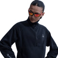 A woman wears the Nike Women’s ACG Wolf Tree 1/2 Zip Top in Black/Anthracite/Summit White, featuring a high collar, her hair pulled back, and reflective red sunglasses. She looks down and to the side against a white background—trail-tested warmth inspired by Nike’s ACG line.