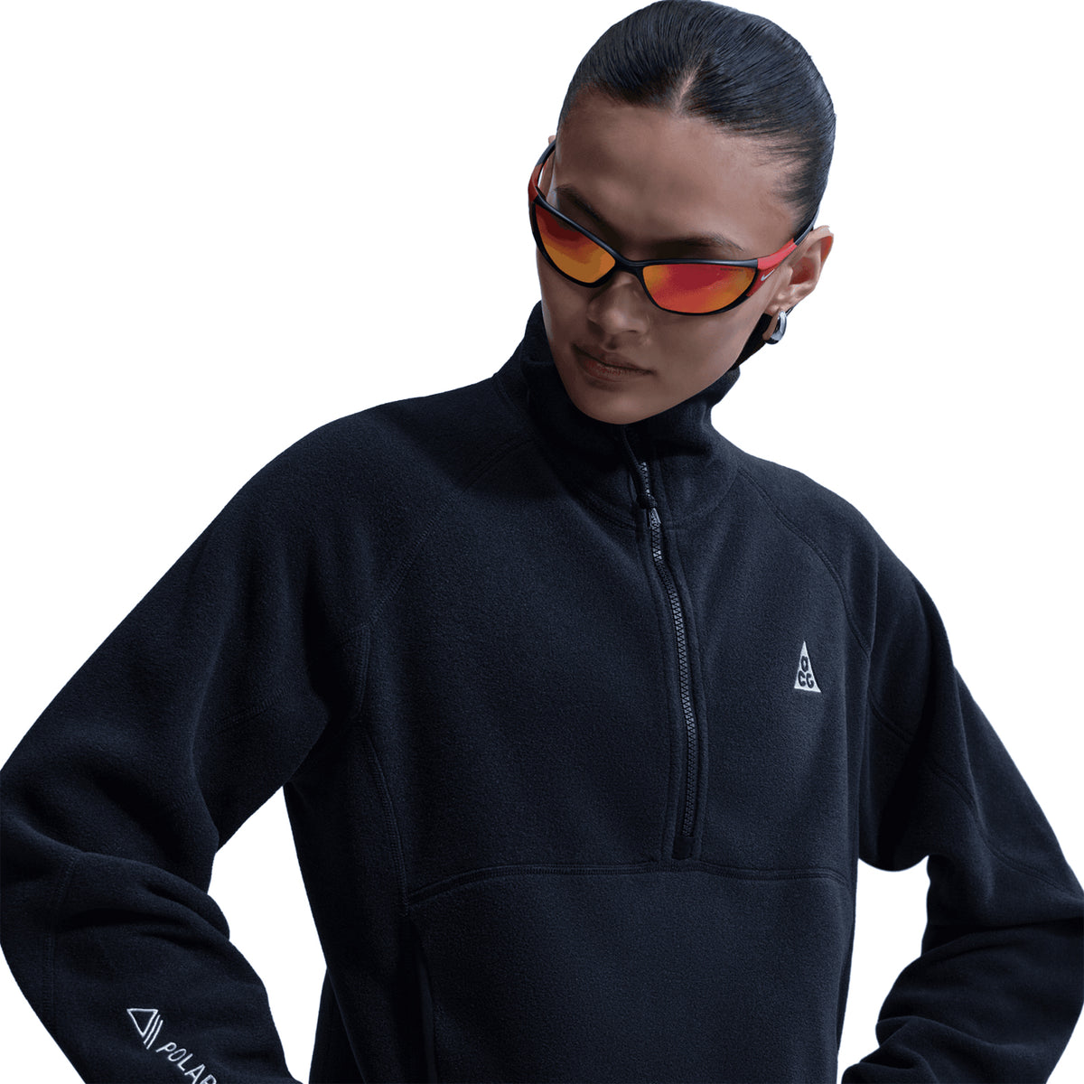A woman wears the Nike Women’s ACG Wolf Tree 1/2 Zip Top in Black/Anthracite/Summit White, featuring a high collar, her hair pulled back, and reflective red sunglasses. She looks down and to the side against a white background—trail-tested warmth inspired by Nike’s ACG line.