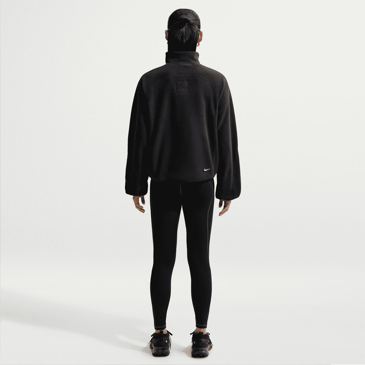 A person stands with their back to the camera against a plain white background, wearing the Nike Women’s ACG Wolf Tree 1/2 Zip Top in Black/Anthracite/Summit White, paired with black leggings, black shoes, and a black cap.