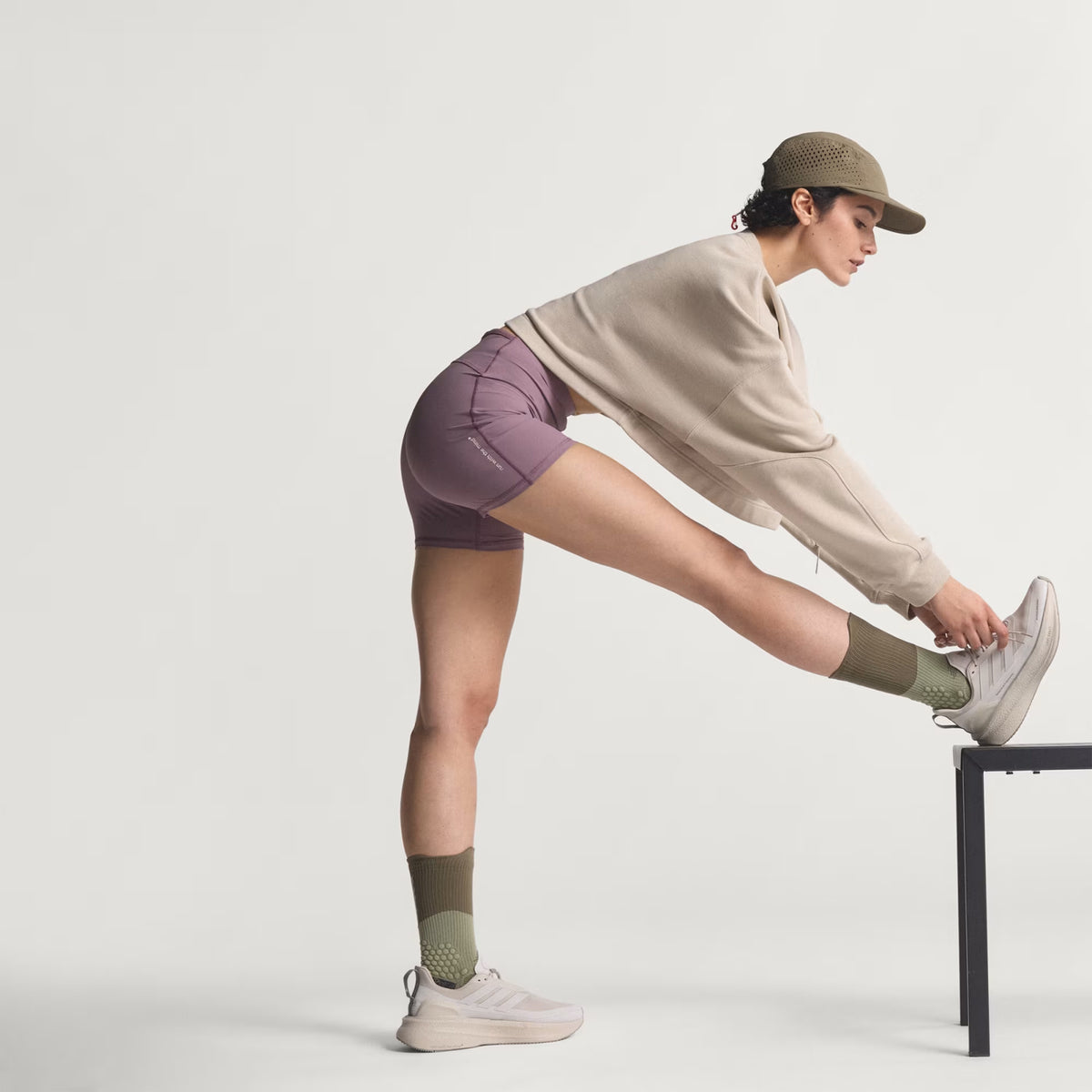 Woman in a beige sweatshirt, purple shorts, green socks, sneakers, and a cap stretches her leg on a bench against a plain background.
