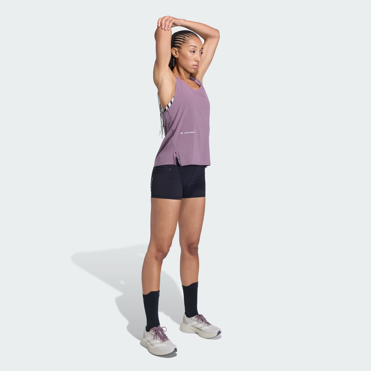 A woman in athletic wear stands stretching her arm overhead. She wears a purple tank top, black shorts, black socks, and white sneakers. She faces slightly to the left against a plain light background.