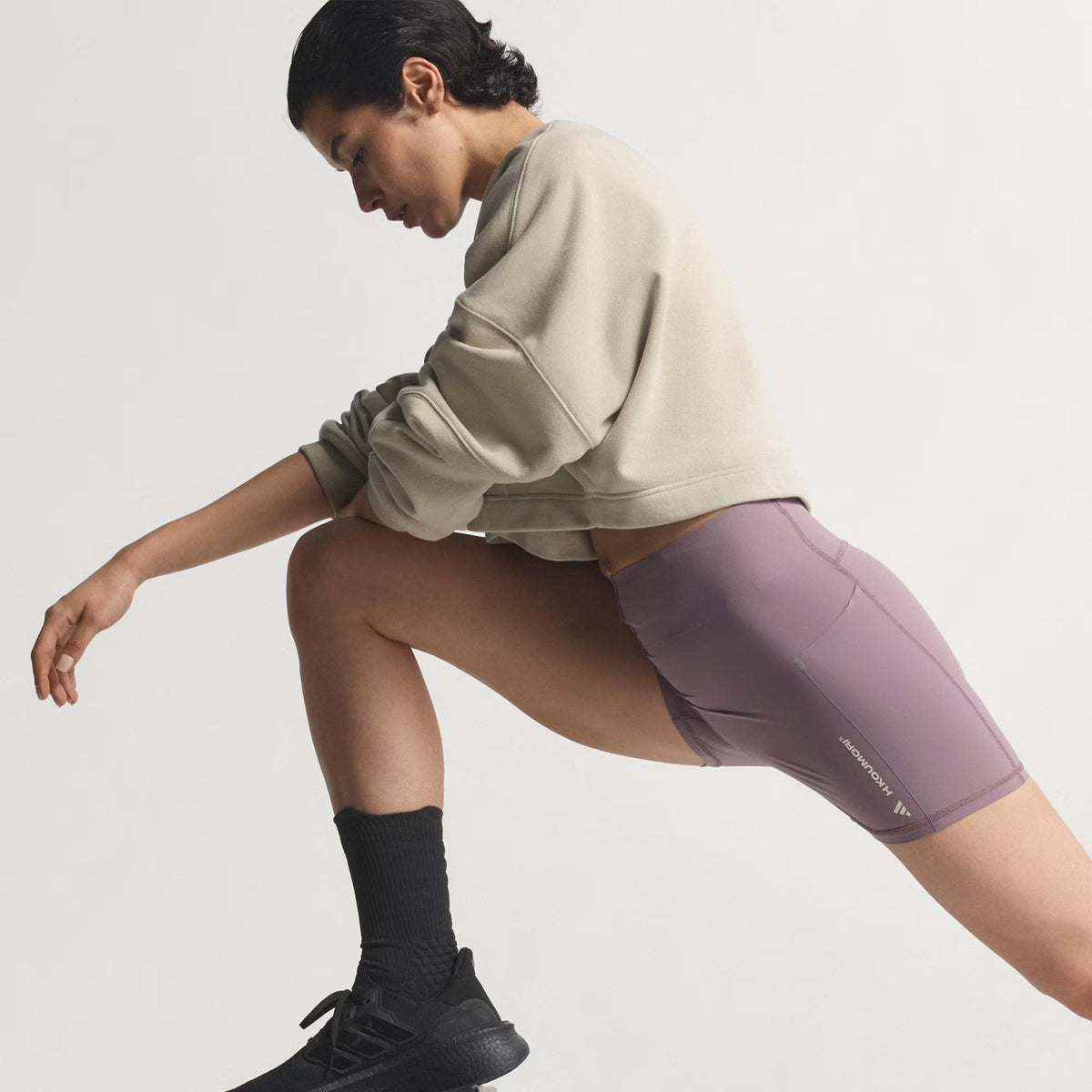 A person in a beige sweatshirt and mauve shorts does a deep lunge stretch against a plain background, wearing black socks and running shoes.